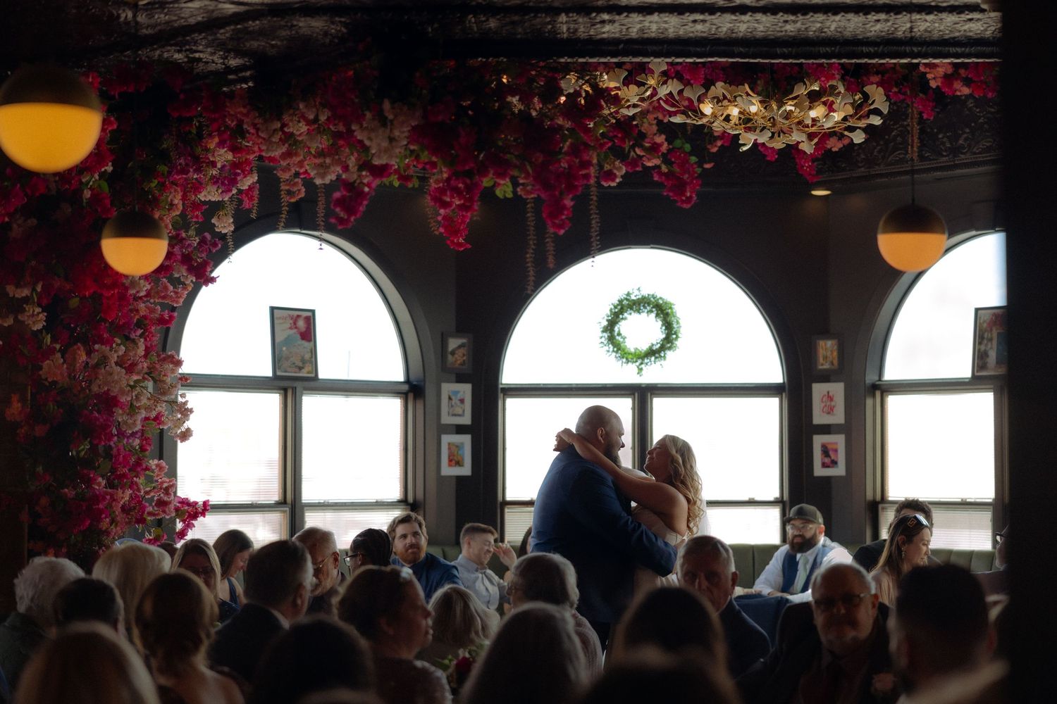 Couple shares a kiss at elegant wedding reception adorned with lush red floral ceiling decor and arched windows.