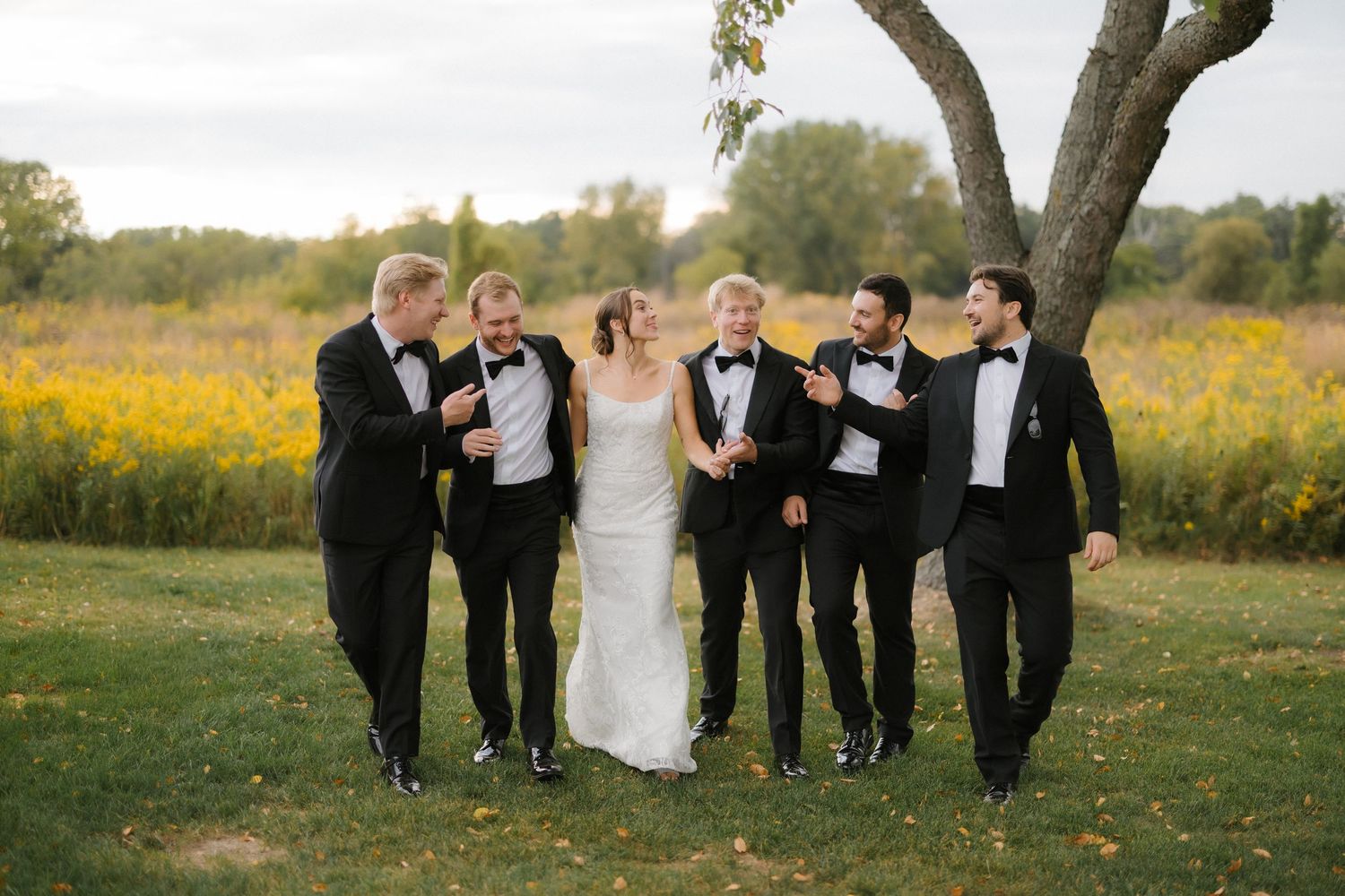 Bride in elegant white gown walking and laughing with groomsmen in black tuxedos outdoors.