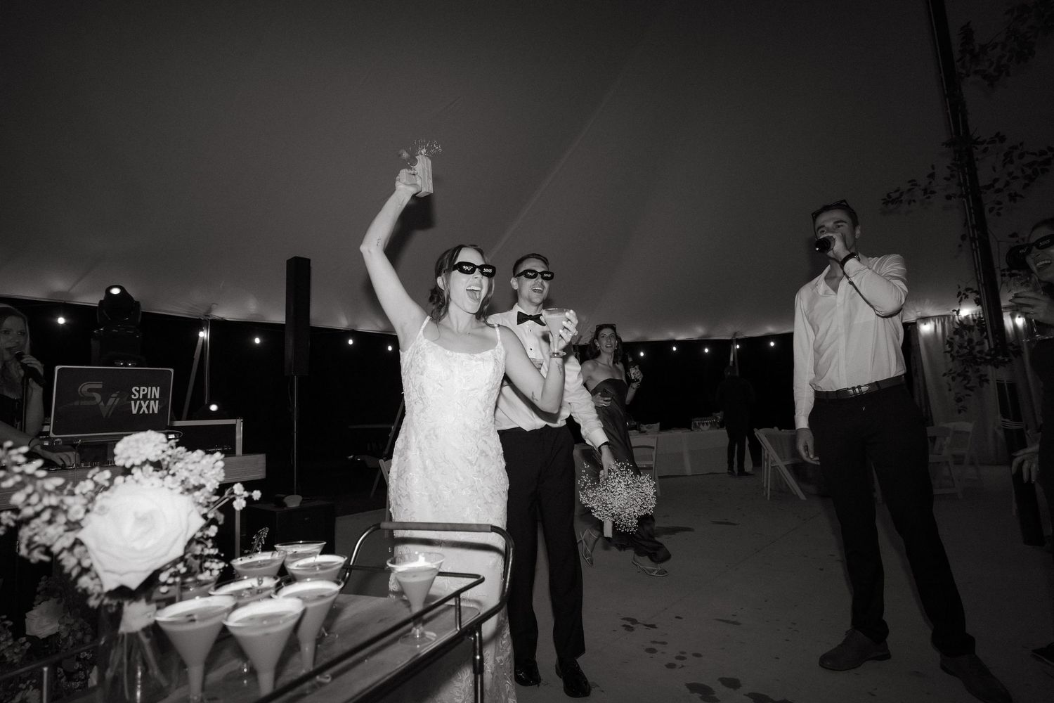 Black and white photo of bride and groom in sunglasses celebrating at their wedding reception table.