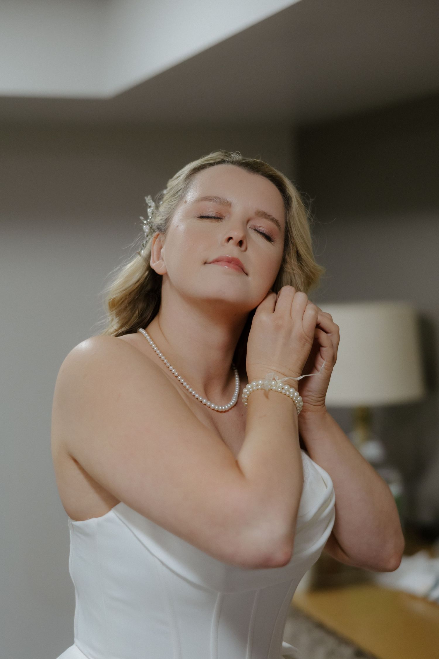 Bride in white strapless gown wearing pearl necklace and bracelet, eyes closed, touching her neck gracefully.
