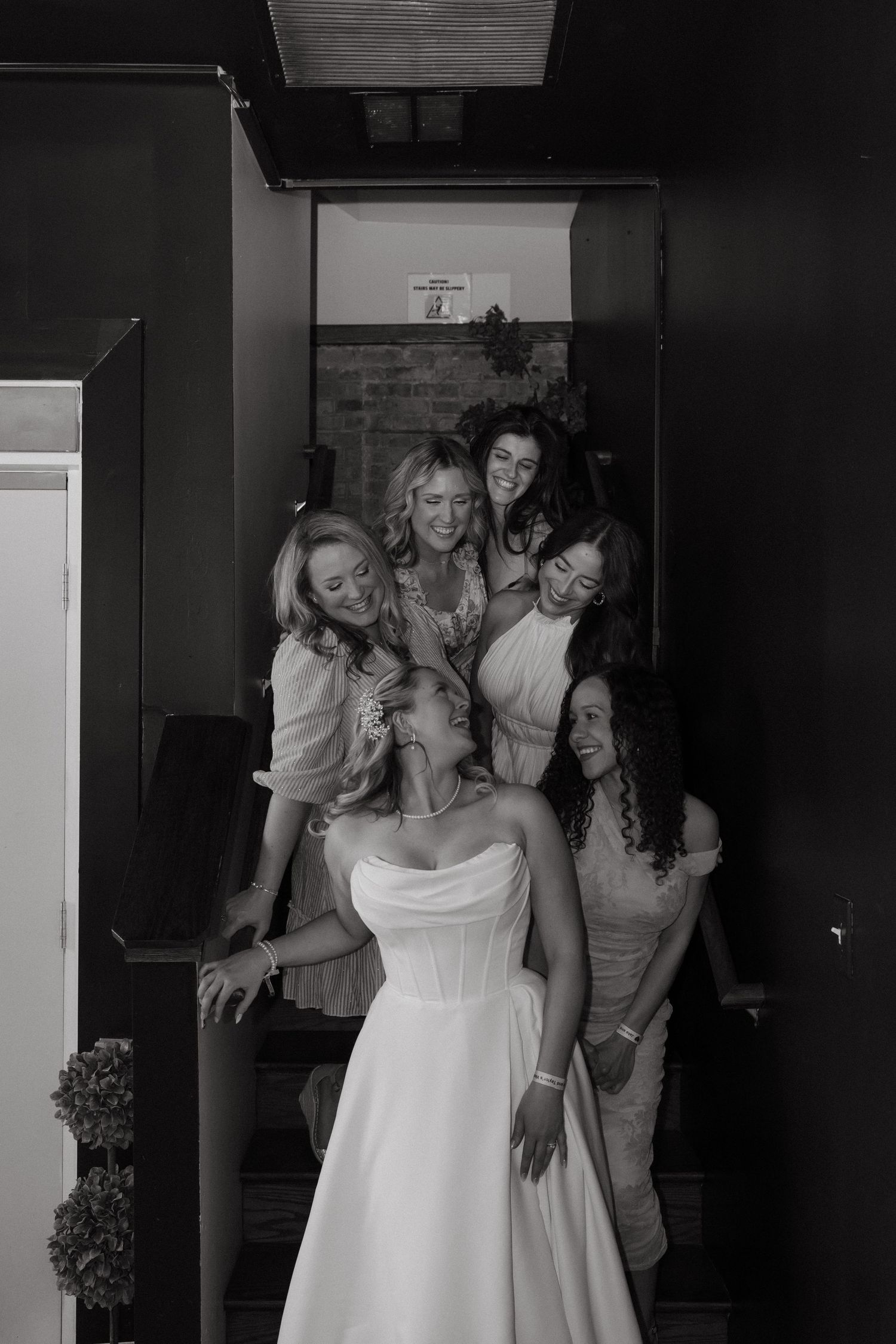 Black and white photo of a bride in a strapless gown surrounded by smiling bridesmaids on a staircase.