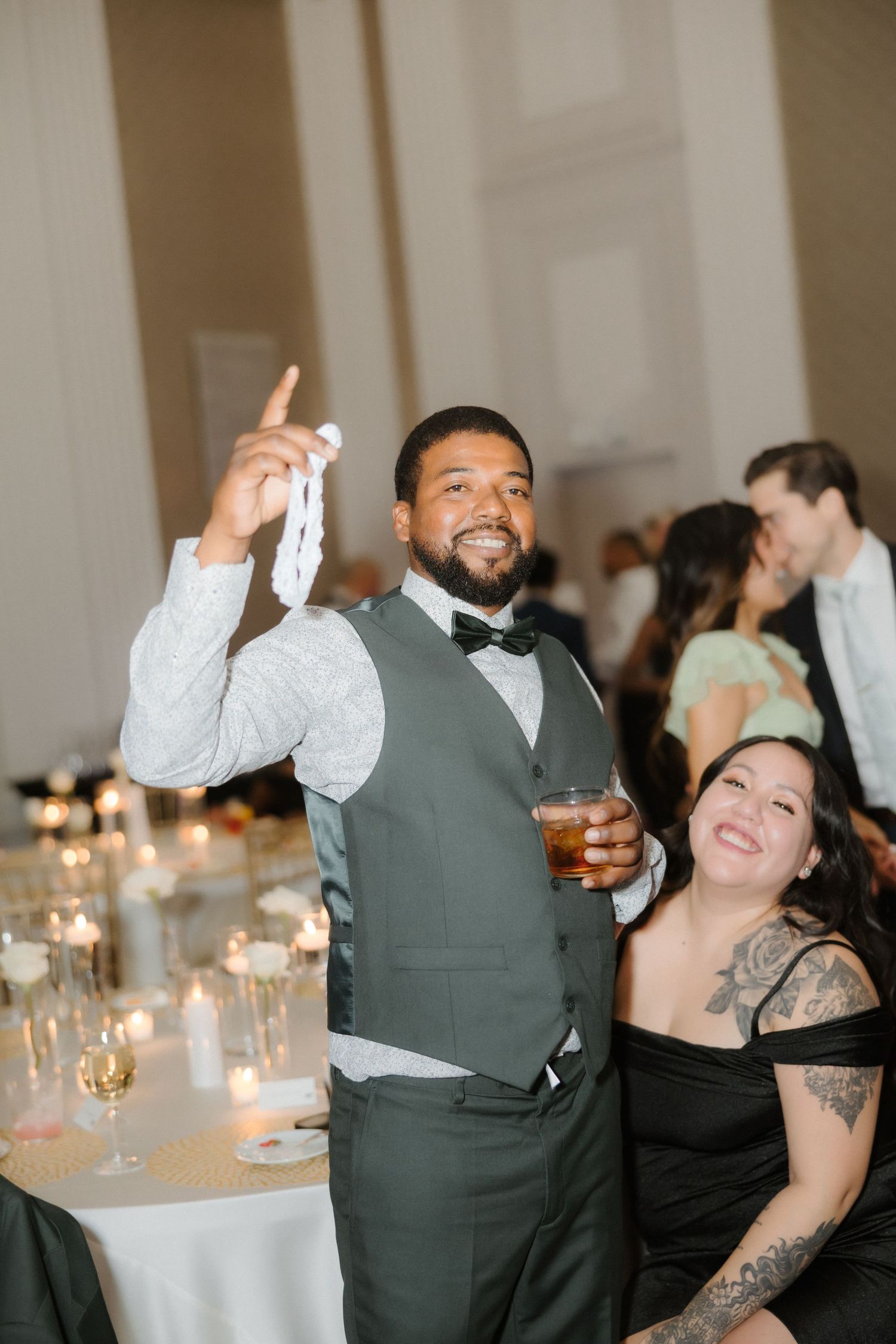 A groomsman in a green vest and bow tie holds a drink and white ribbon aloft, smiling at a wedding reception.