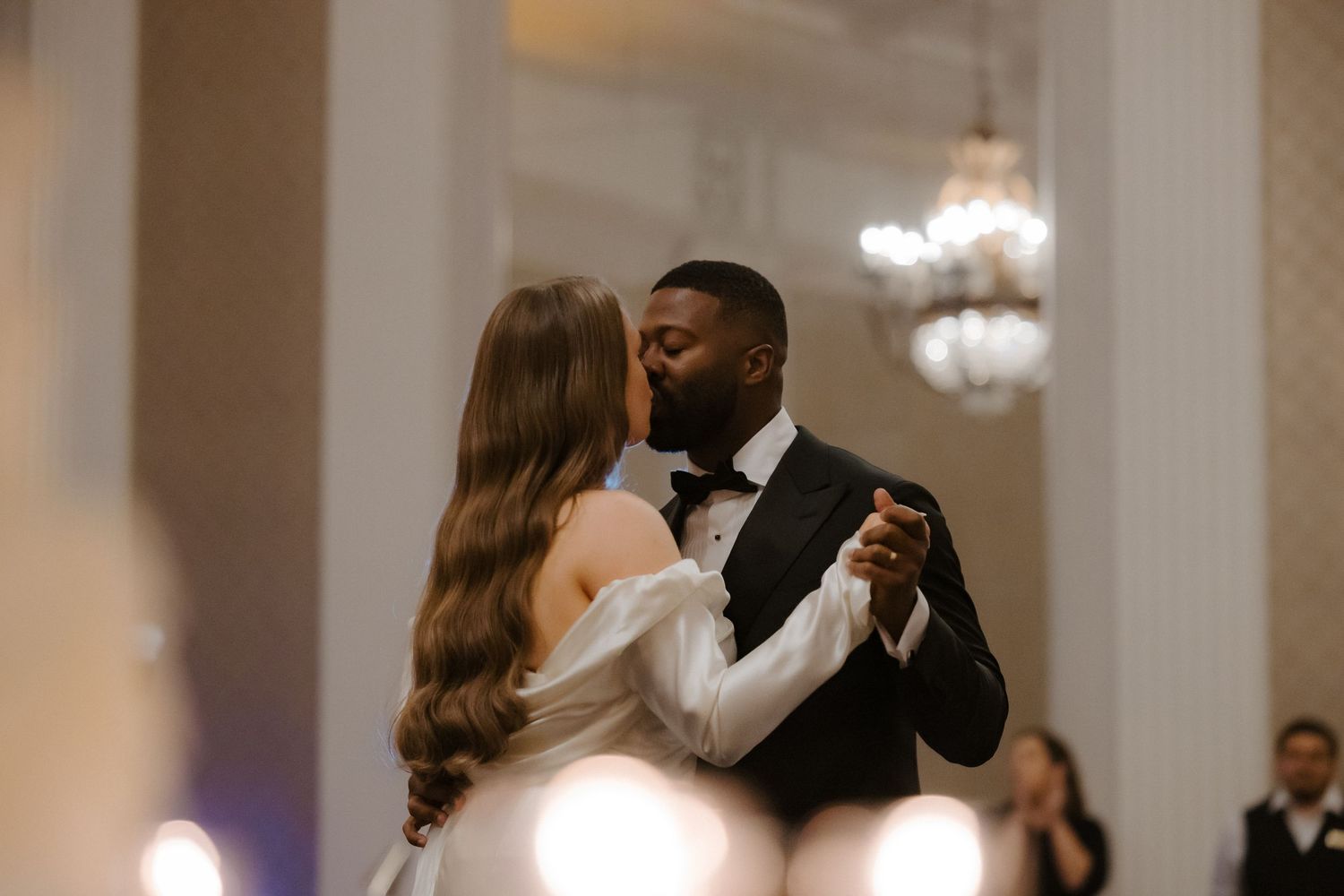 A bride and groom share a romantic first dance kiss in an elegant ballroom with chandeliers and white columns.
