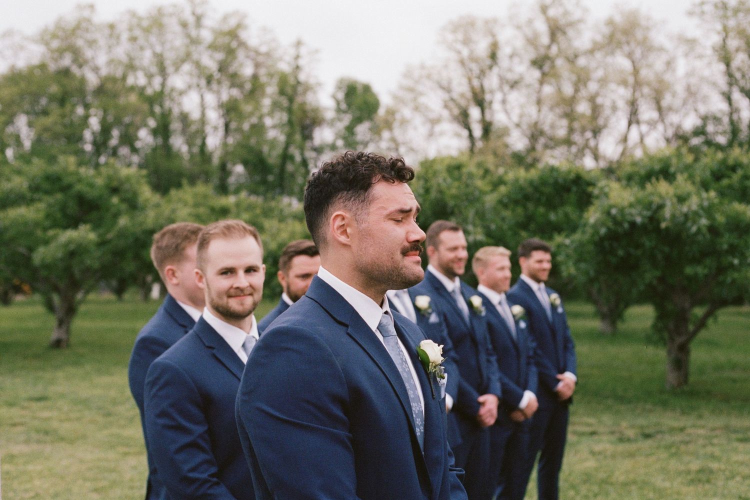 Groom and groomsmen in navy suits with white boutonnieres standing outdoors in an orchard during a wedding ceremony.