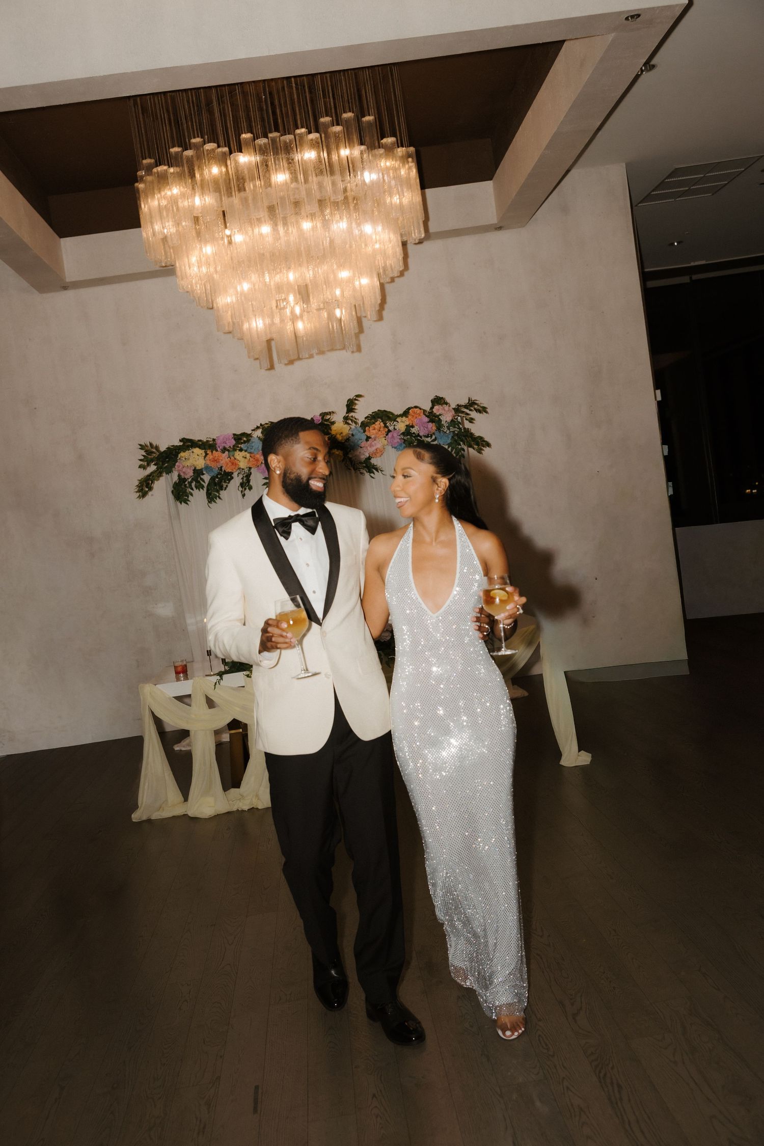 Elegant couple in wedding attire walking under chandelier at reception venue.