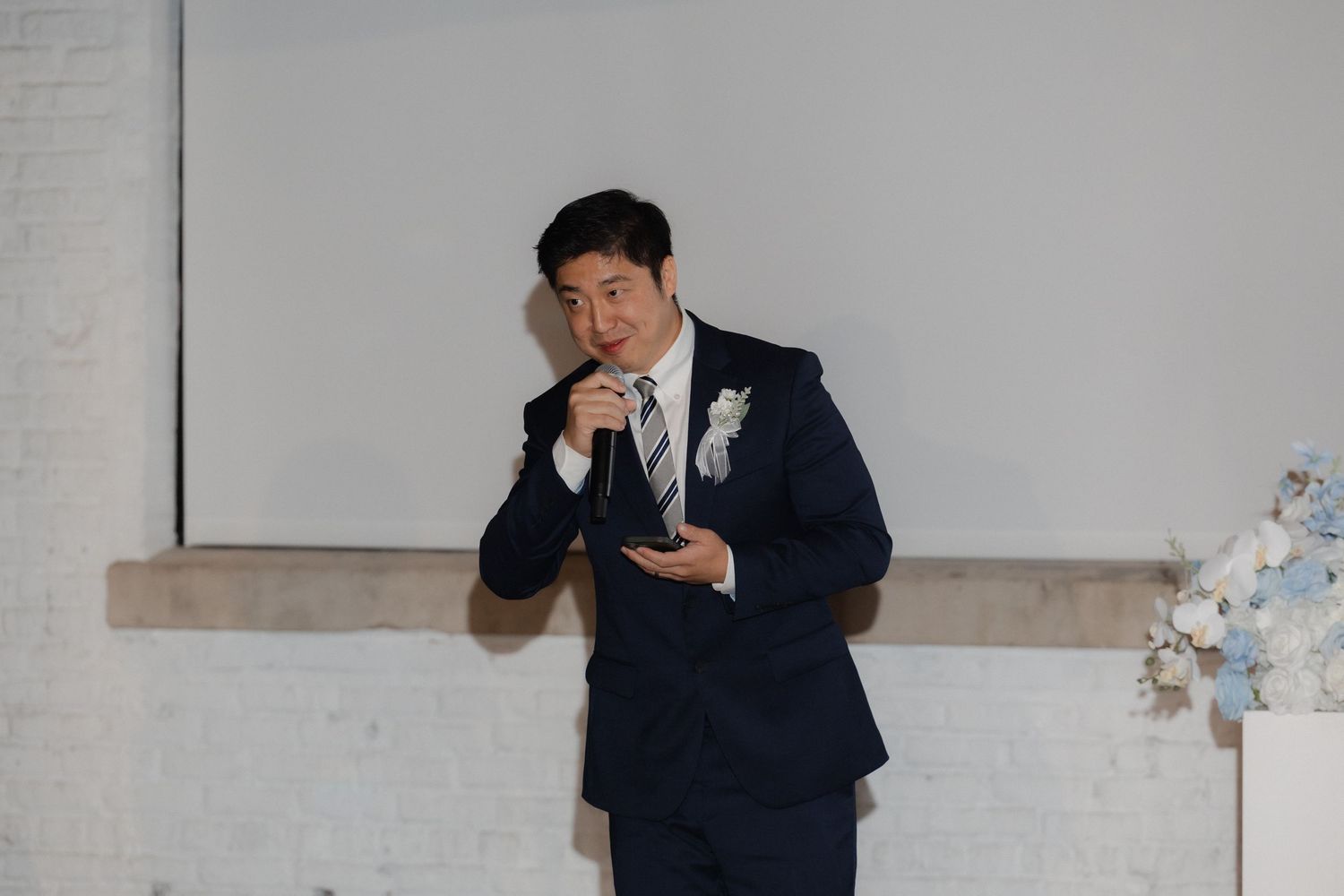 Man in navy suit speaks into microphone at wedding reception with floral arrangement nearby.