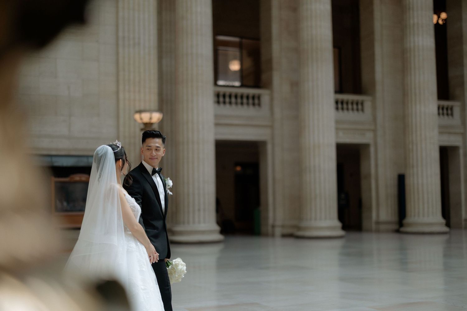 A bride and groom share a romantic moment among grand columns in an elegant wedding venue.