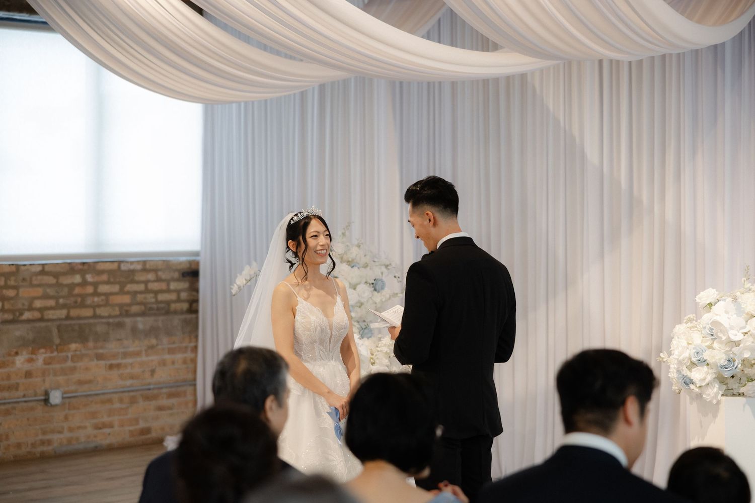 A bride and groom exchange vows at an elegant wedding ceremony with white floral arrangements and draped fabric.