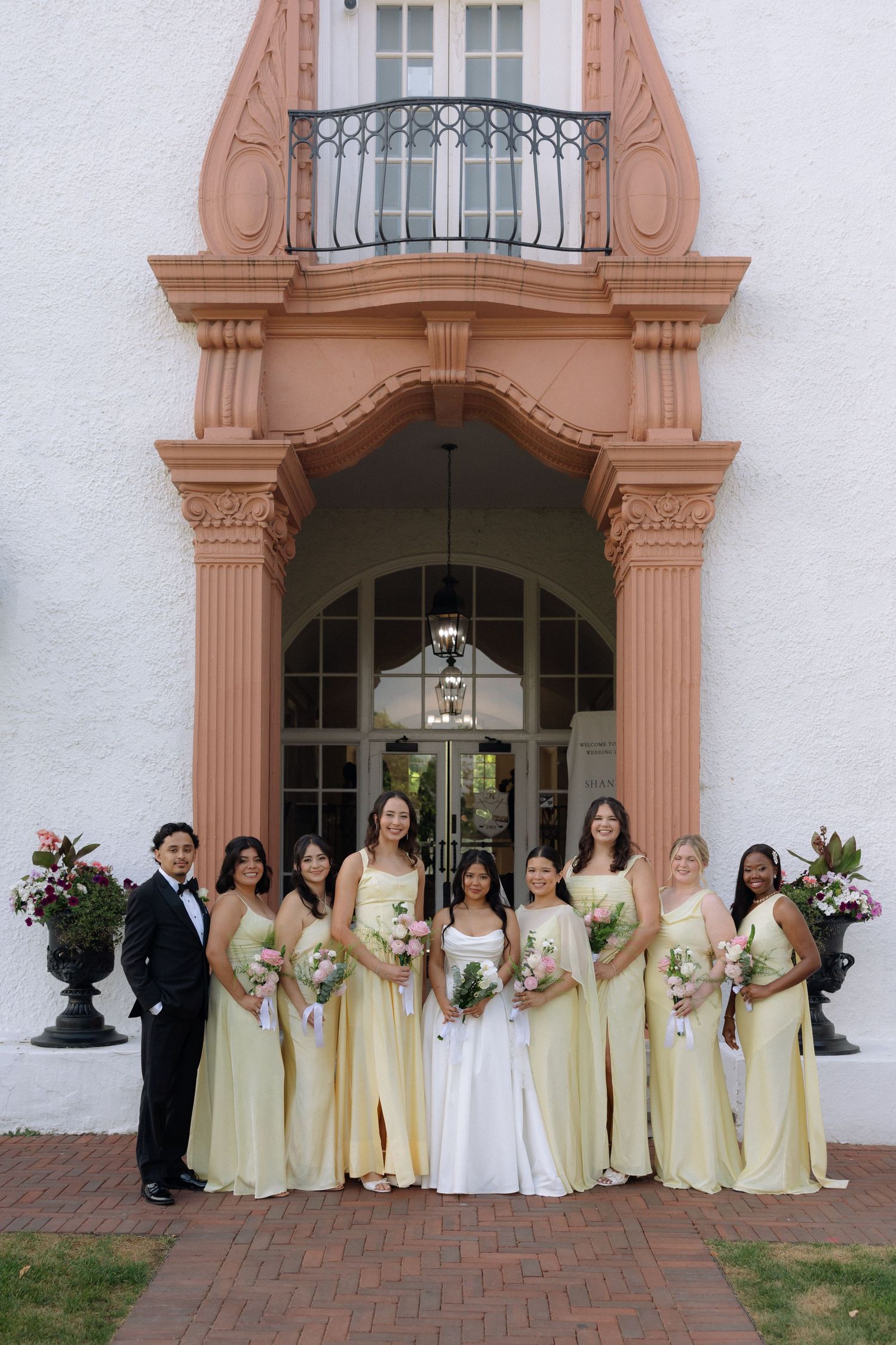 Bride in white gown with bridesmaids in yellow dresses and groomsman in tuxedo posing outside elegant venue.