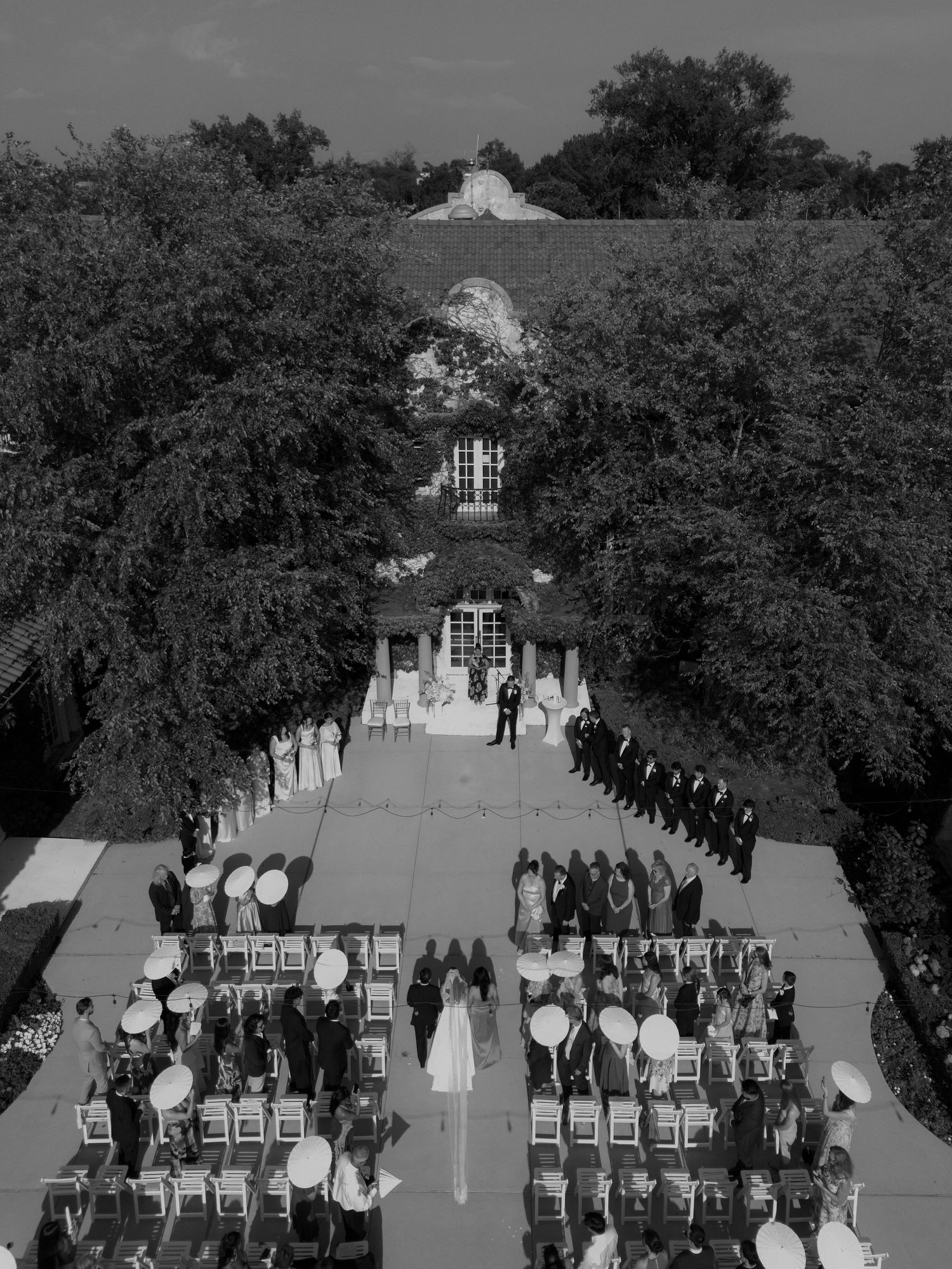Aerial black and white drone shot of an elegant outdoor wedding ceremony with guests seated in rows along a tree-lined aisle.