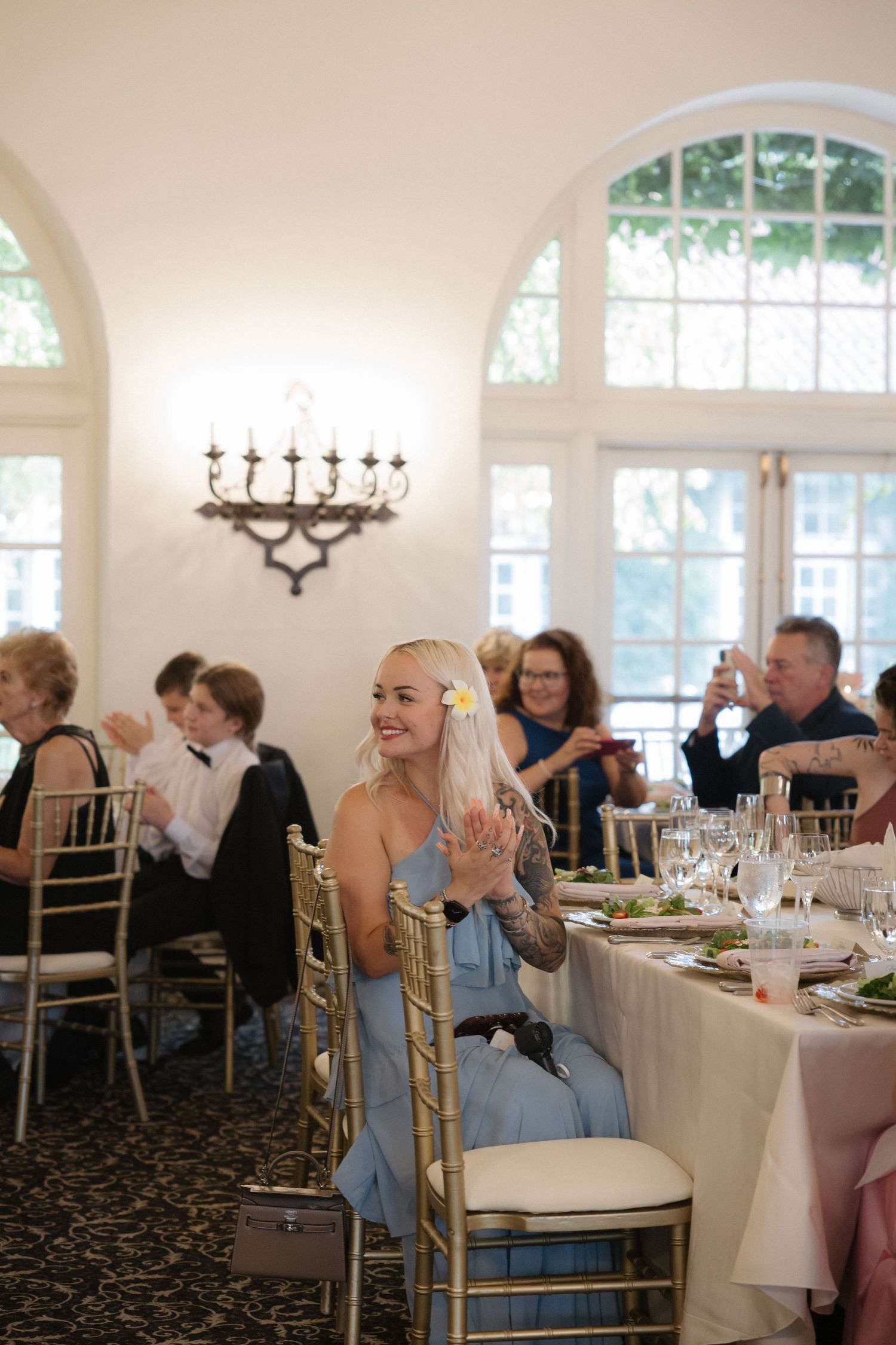 Smiling blonde bridesmaid in blue dress clapping at wedding reception in elegant white venue with arched windows.
