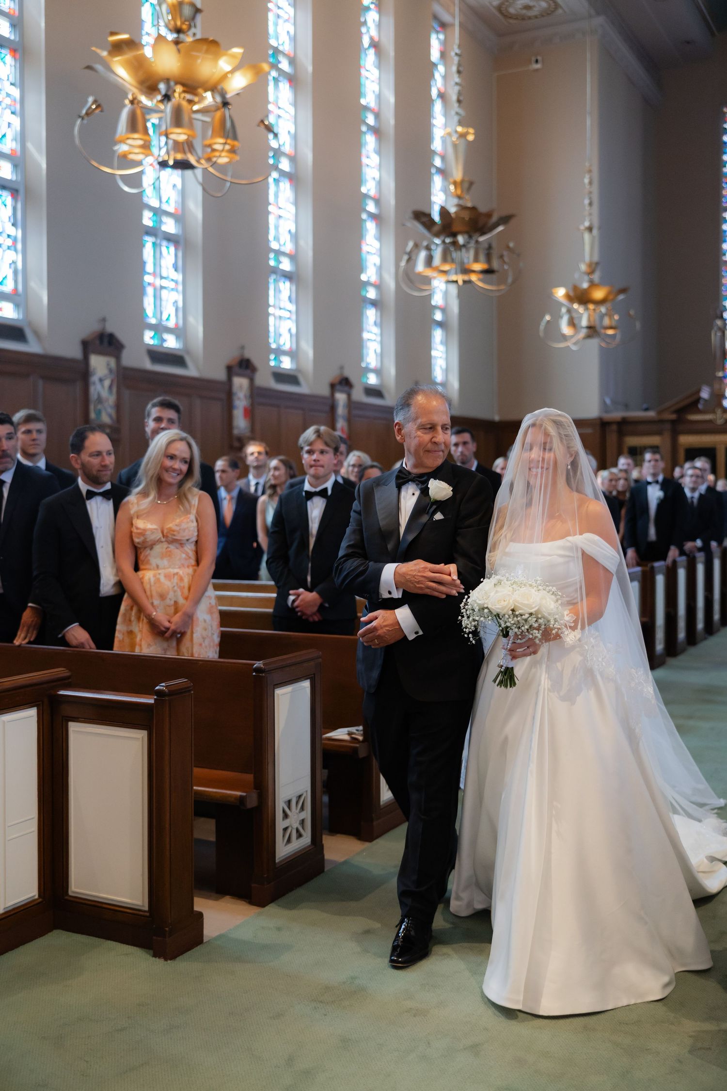 Bride and father walk down ornate church aisle with stained glass windows during ceremony.