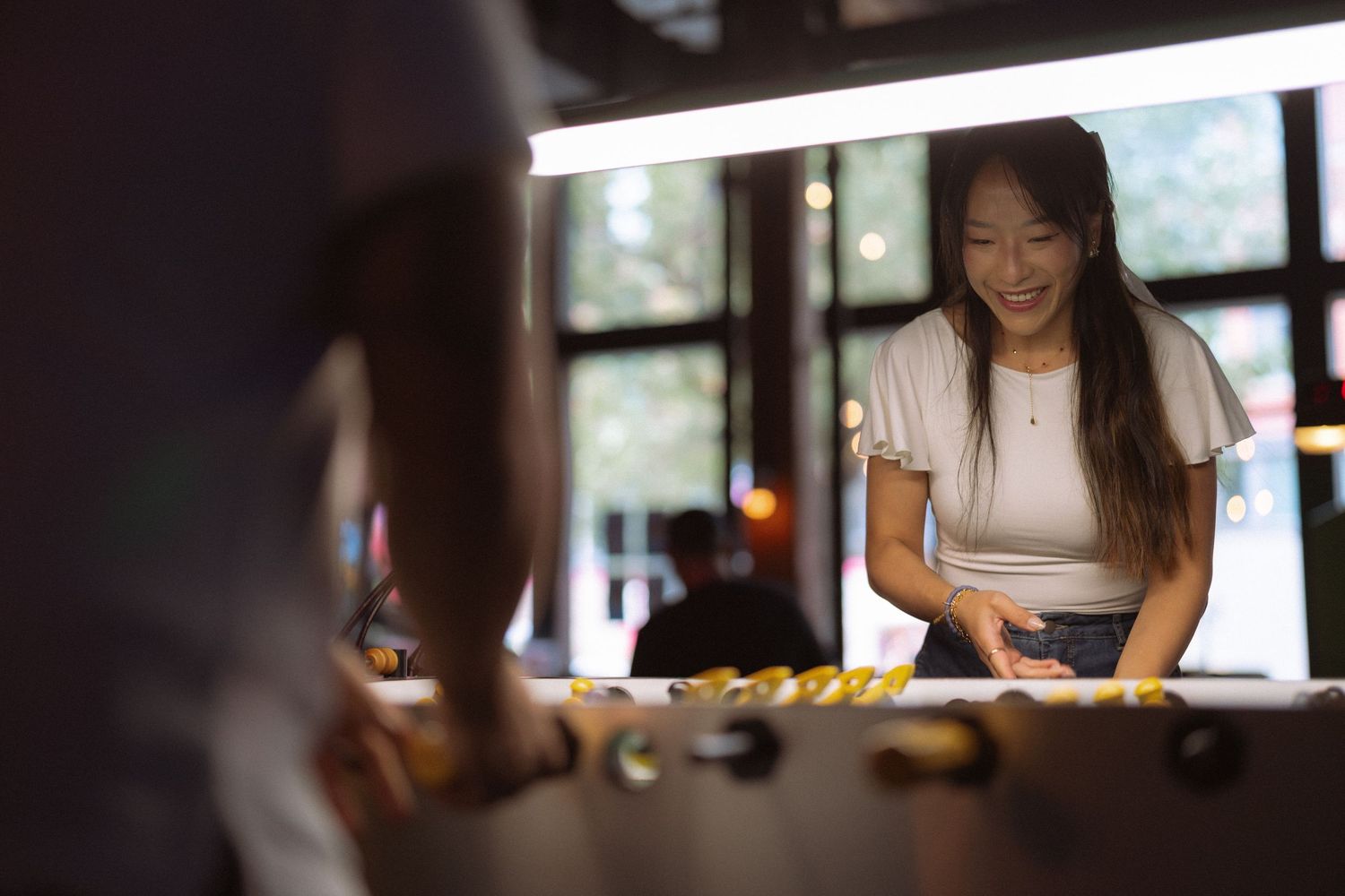 Smiling Asian woman playing foosball at a game table in a modern bar or entertainment venue.