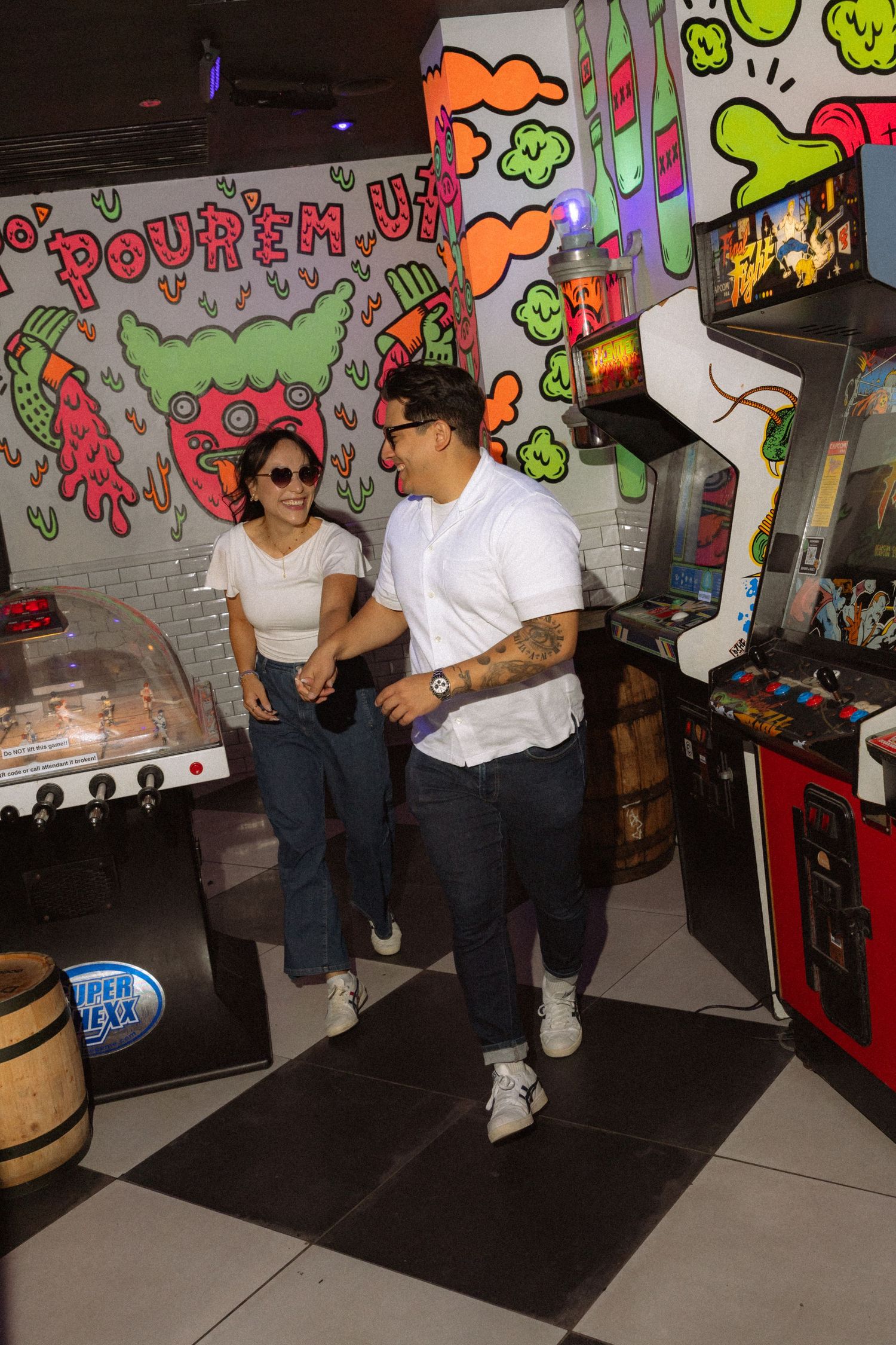 Couple in white tees and jeans enjoying a colorful retro arcade bar with vintage games and vibrant graffiti wall art.