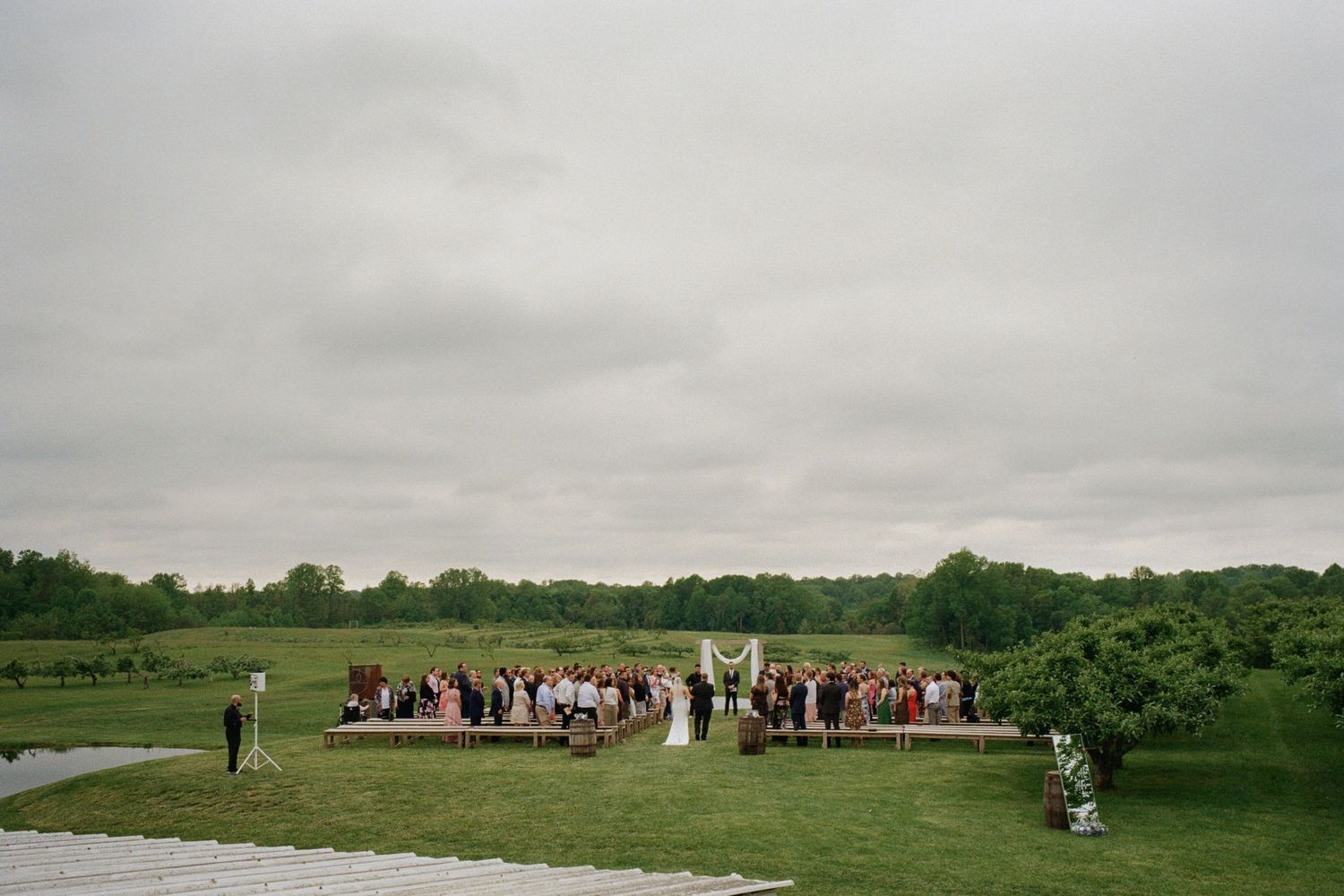 Outdoor wedding ceremony on green lawn with guests gathered under arch, overcast sky, rural countryside backdrop.