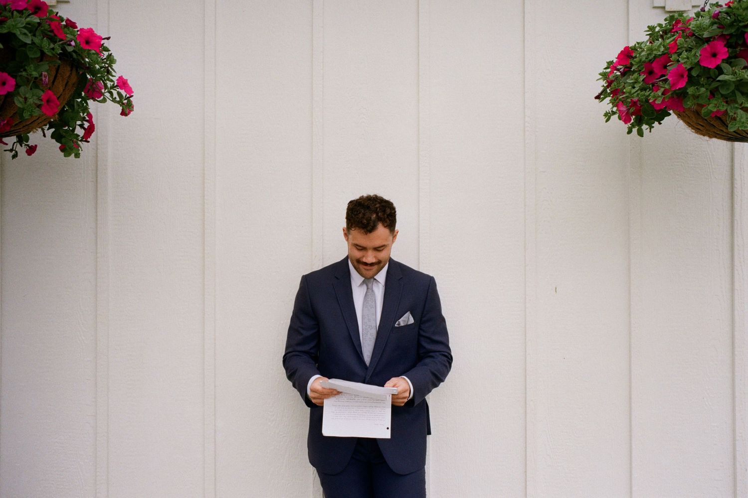 Groom in navy suit reads vows paper outdoors, flanked by pink hanging flower baskets on white wall.