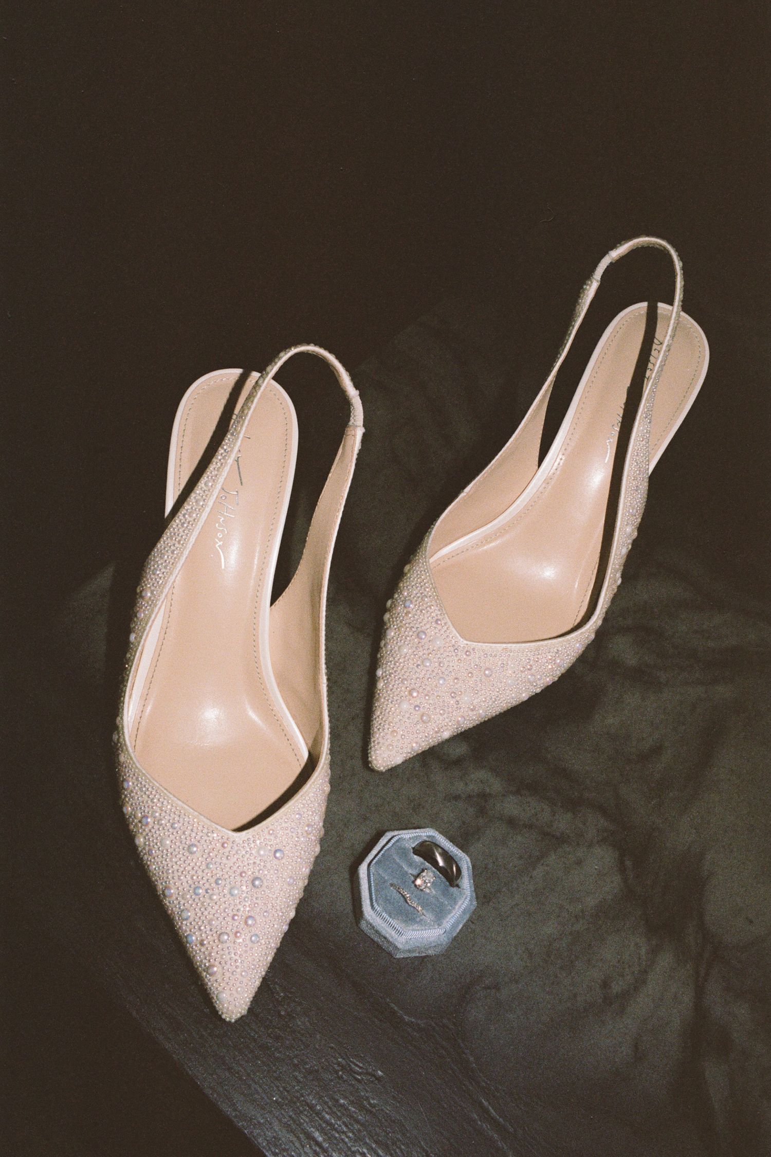 Elegant nude crystal-embellished slingback heels displayed beside a small blue hexagonal ring box on dark surface.