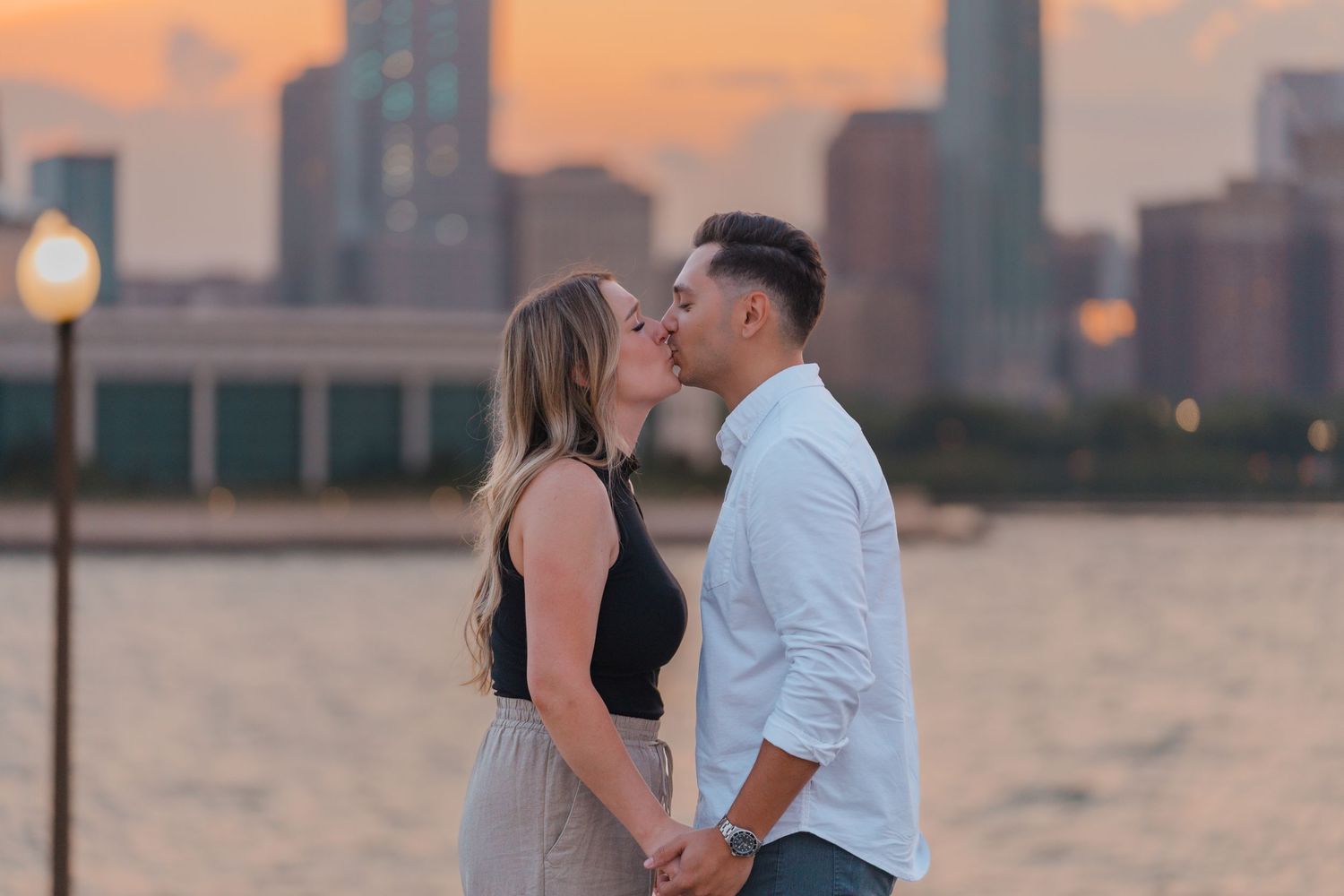 Newly engaged couple kissing while holding hands with a glowing city skyline at dusk.