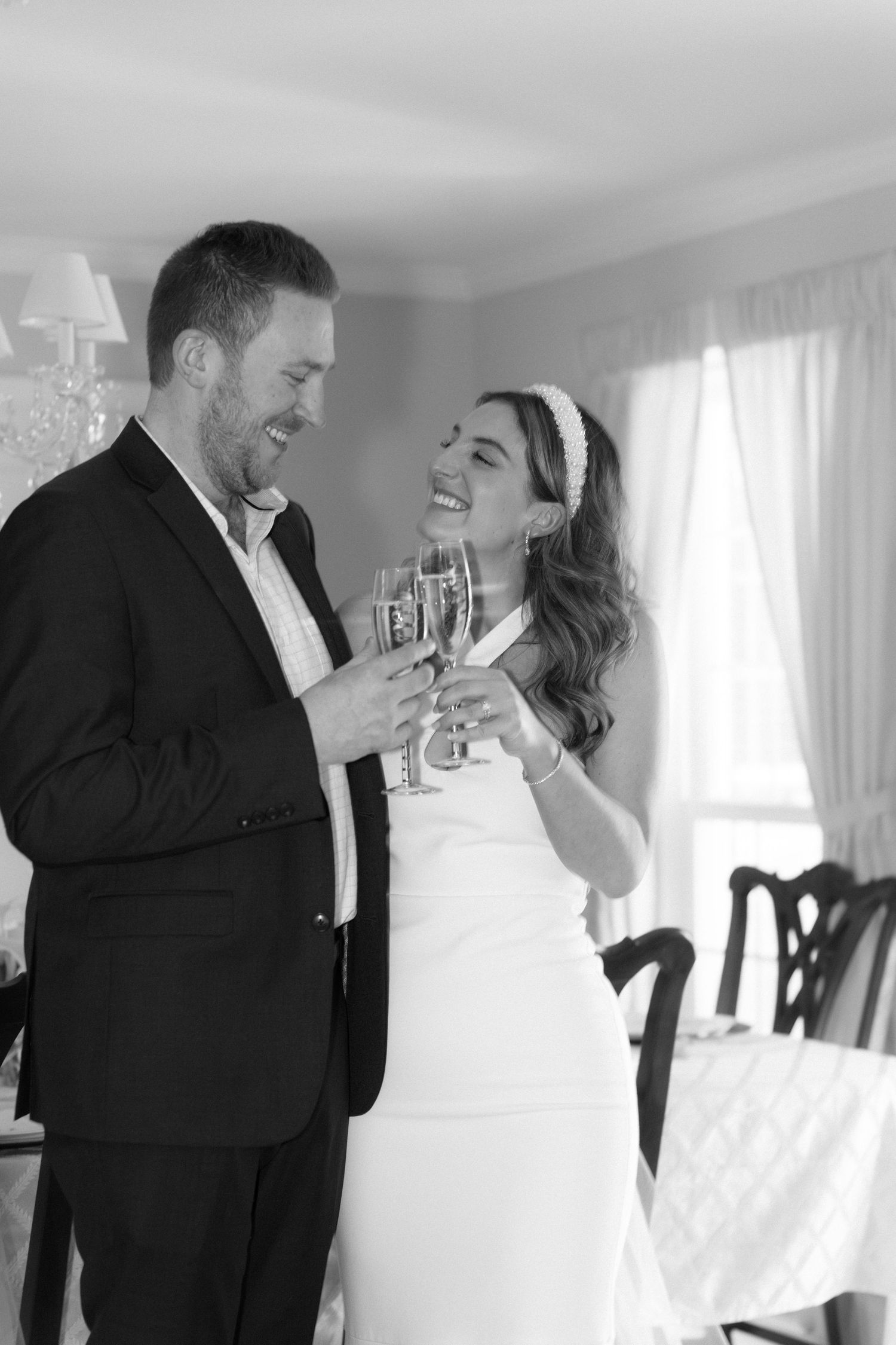 Black and white photo of smiling bride and groom clinking champagne glasses at their wedding celebration.