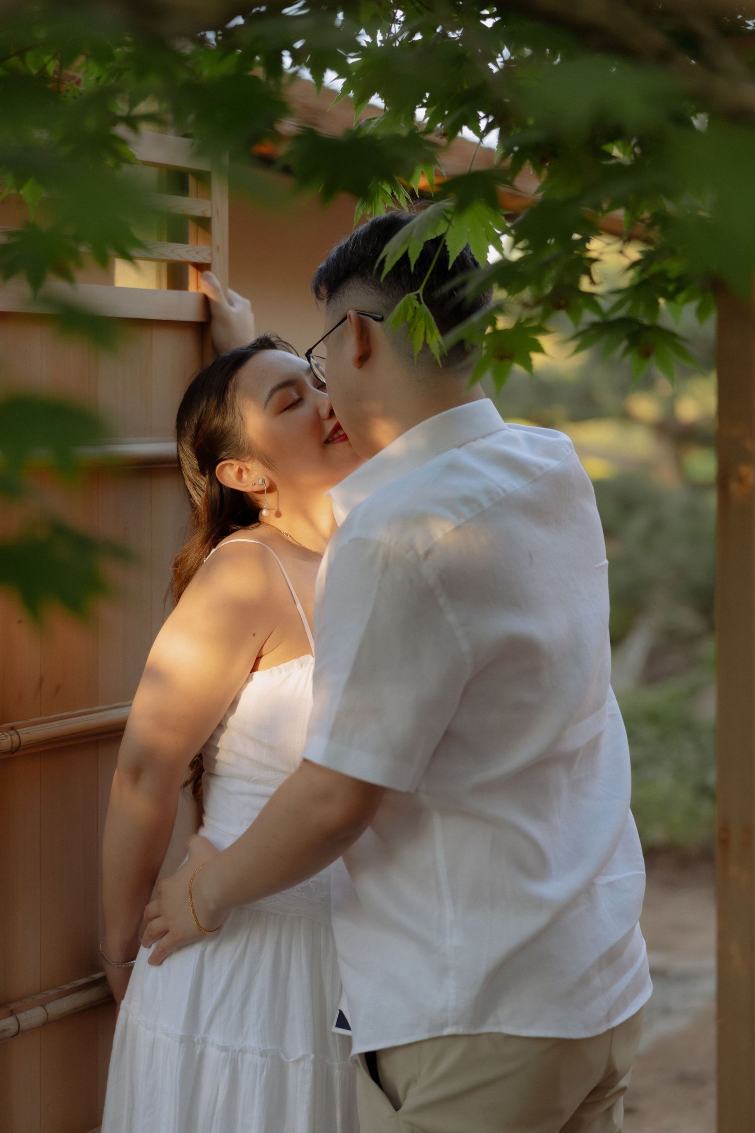 A couple sharing a romantic kiss outdoors, framed by green maple leaves, the woman in a white dress and man in a white shirt.