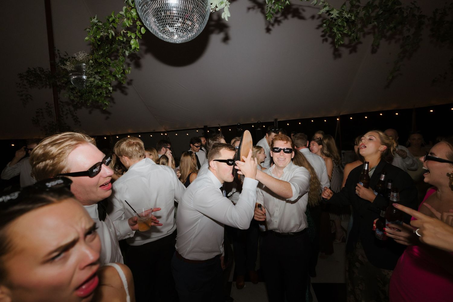 Wedding guests wearing sunglasses high-five and dance energetically under a disco ball at the reception.
