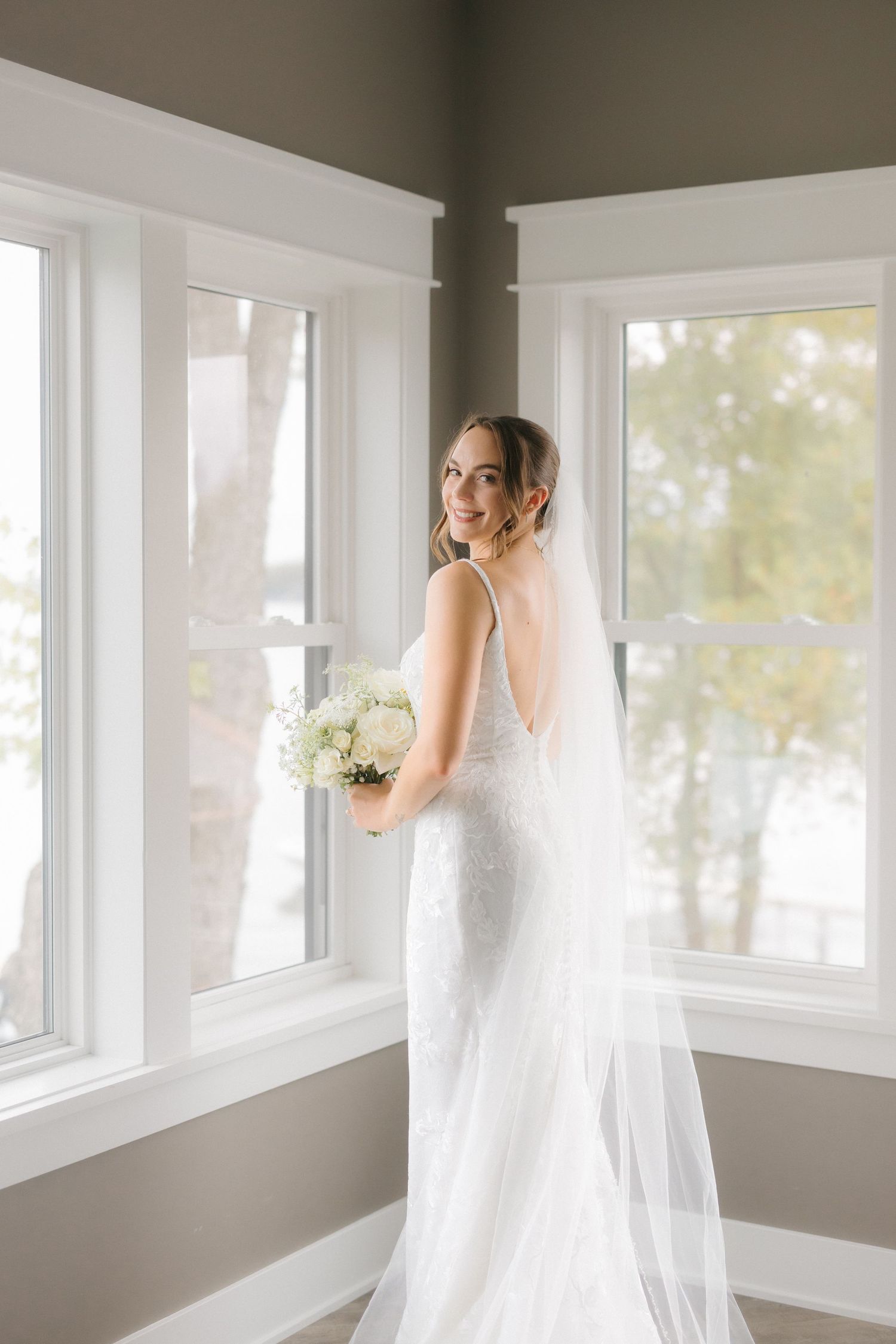 Smiling bride in backless lace wedding dress with flowing veil holding white bouquet, posing by sunlit windows.