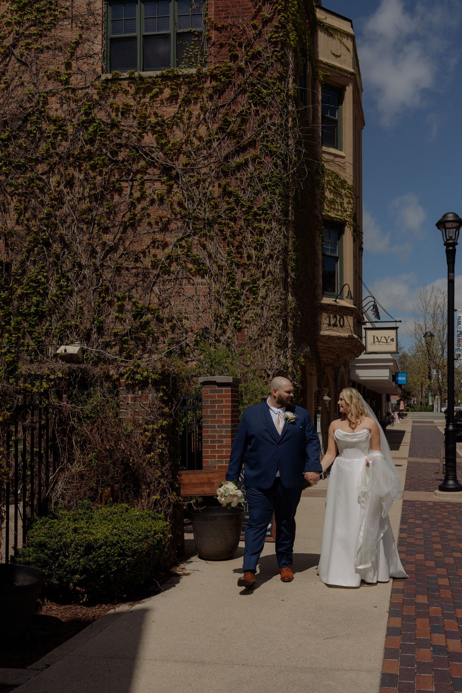 Bride and groom walk hand-in-hand along sidewalk beside ivy-covered brick building on sunny wedding day.