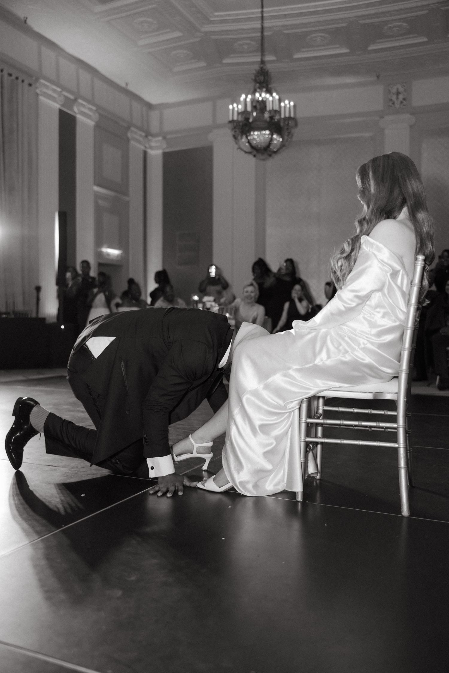 Black and white photo of groom kneeling before bride during garter removal at elegant wedding reception.