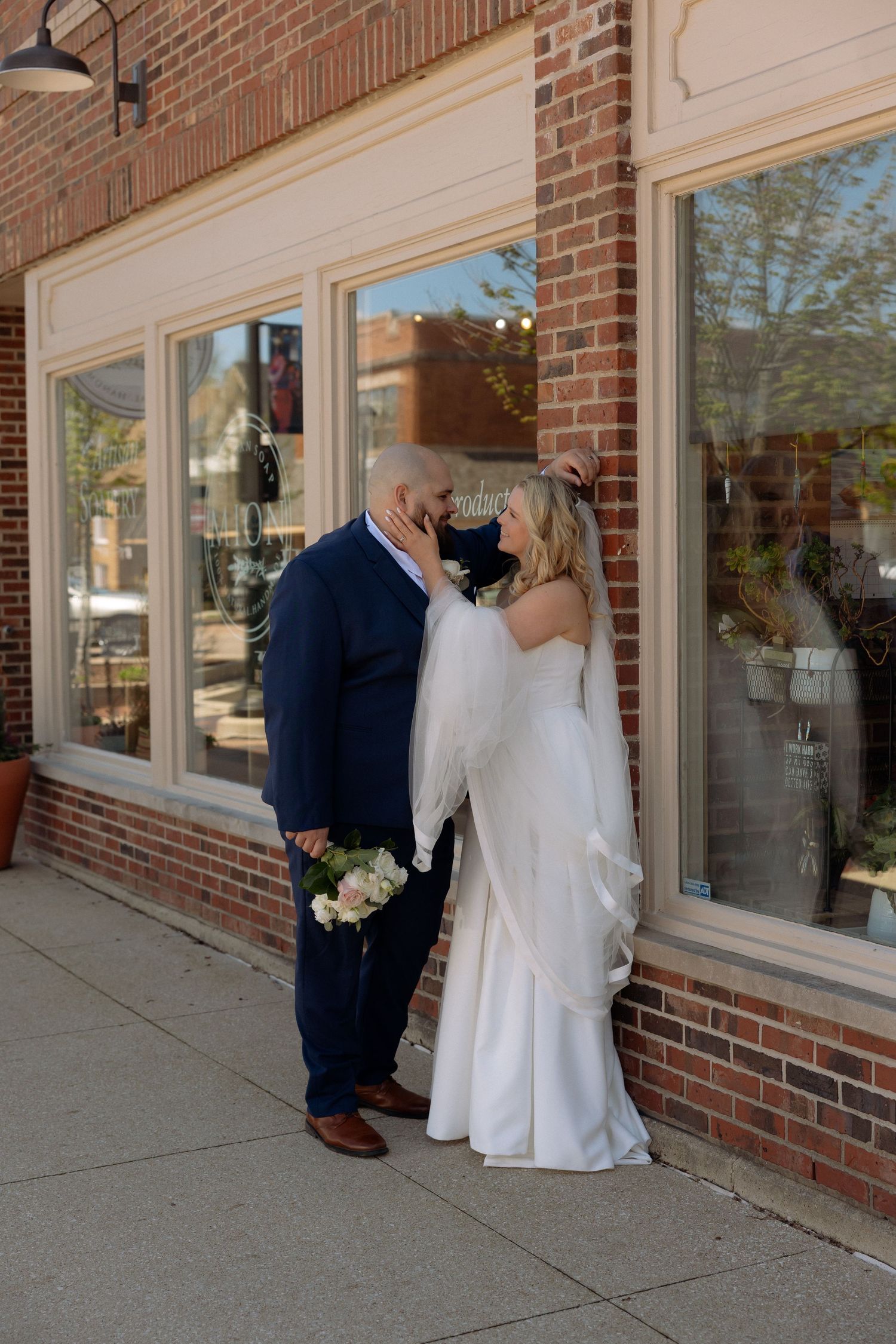 Bride and groom sharing a romantic kiss outside a brick storefront on their wedding day.