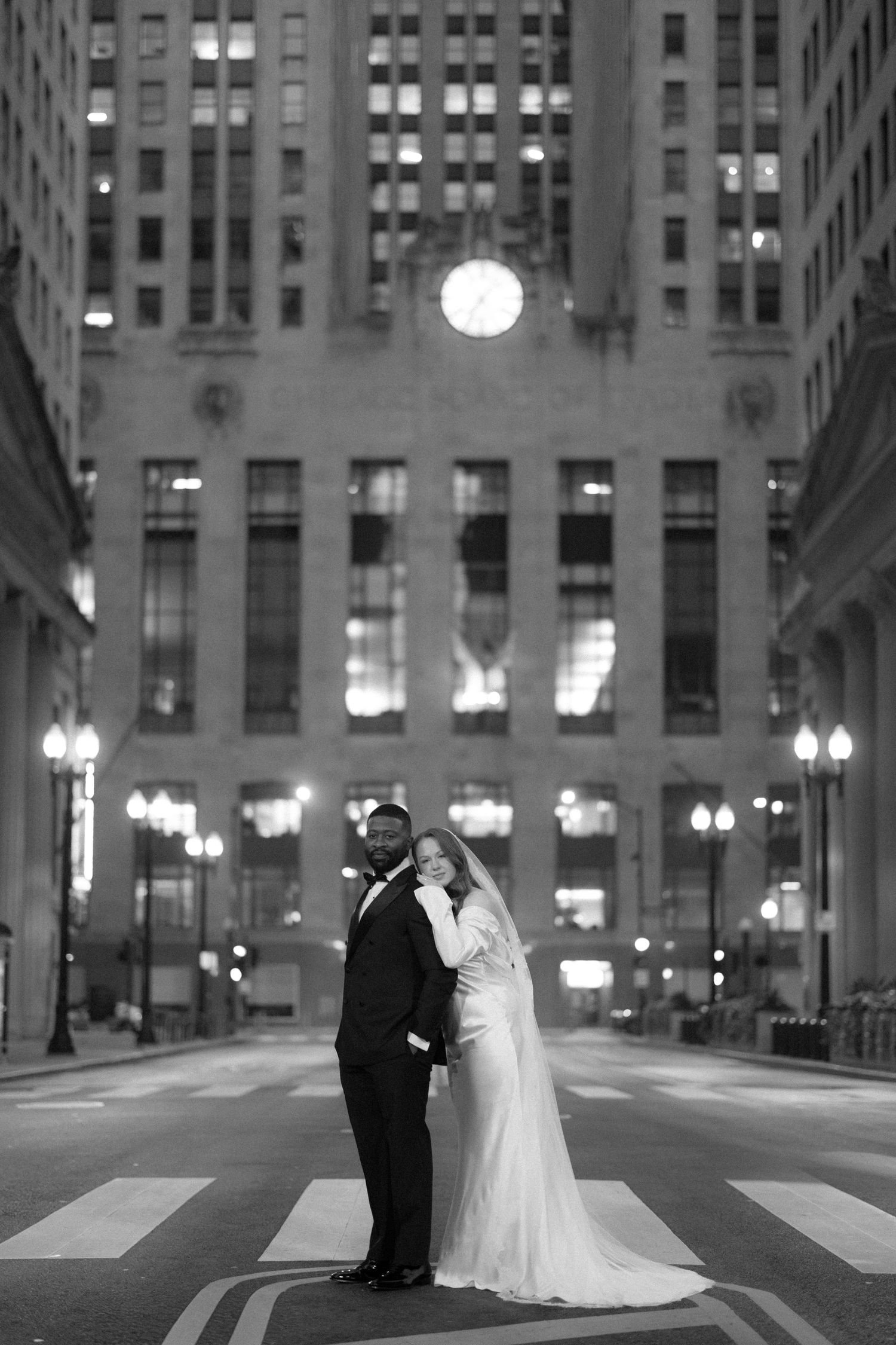 Black and white wedding photo of bride and groom standing on crosswalk in front of illuminated city building at night.