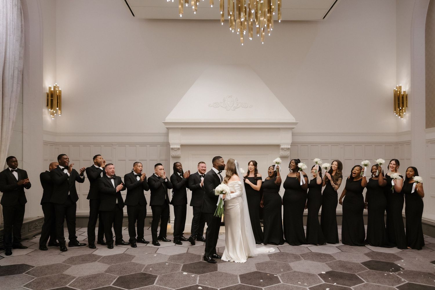 Bride and groom share a kiss surrounded by wedding party in black attire inside an elegant ballroom with chandelier.