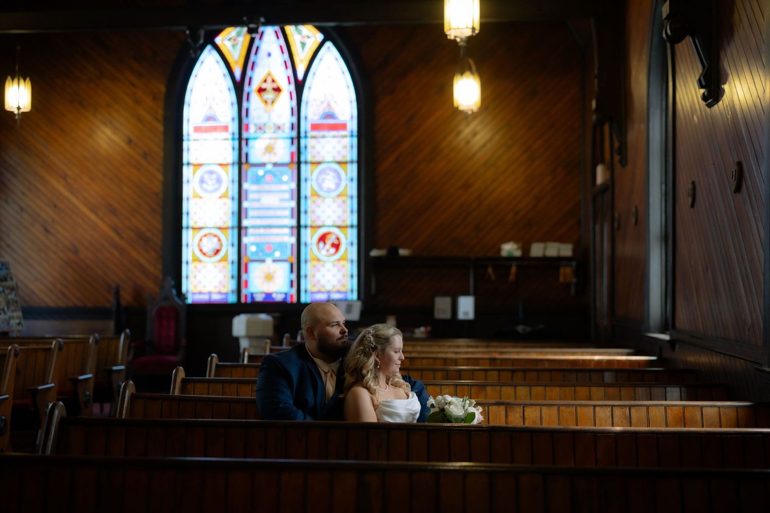 Couple sits together in wooden church pew before colorful stained glass windows during intimate wedding ceremony.