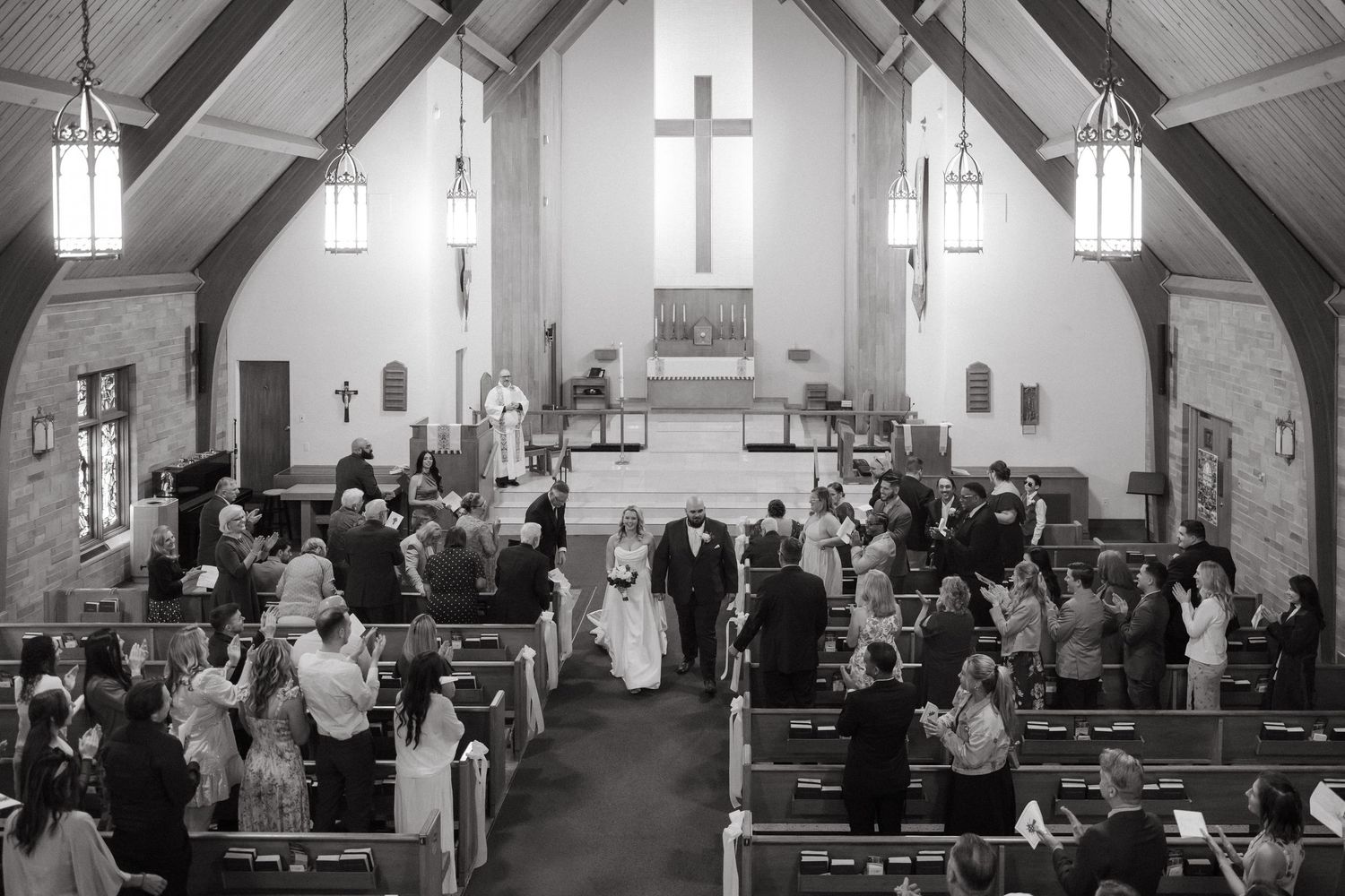 Black and white photo of a wedding ceremony in a church with guests standing in pews as the couple walks down the aisle.