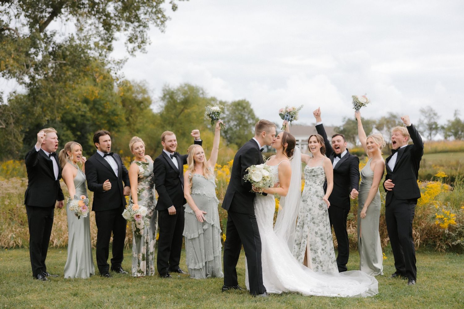 Joyful wedding party cheers as bride and groom share a kiss surrounded by golden autumn wildflowers and greenery.