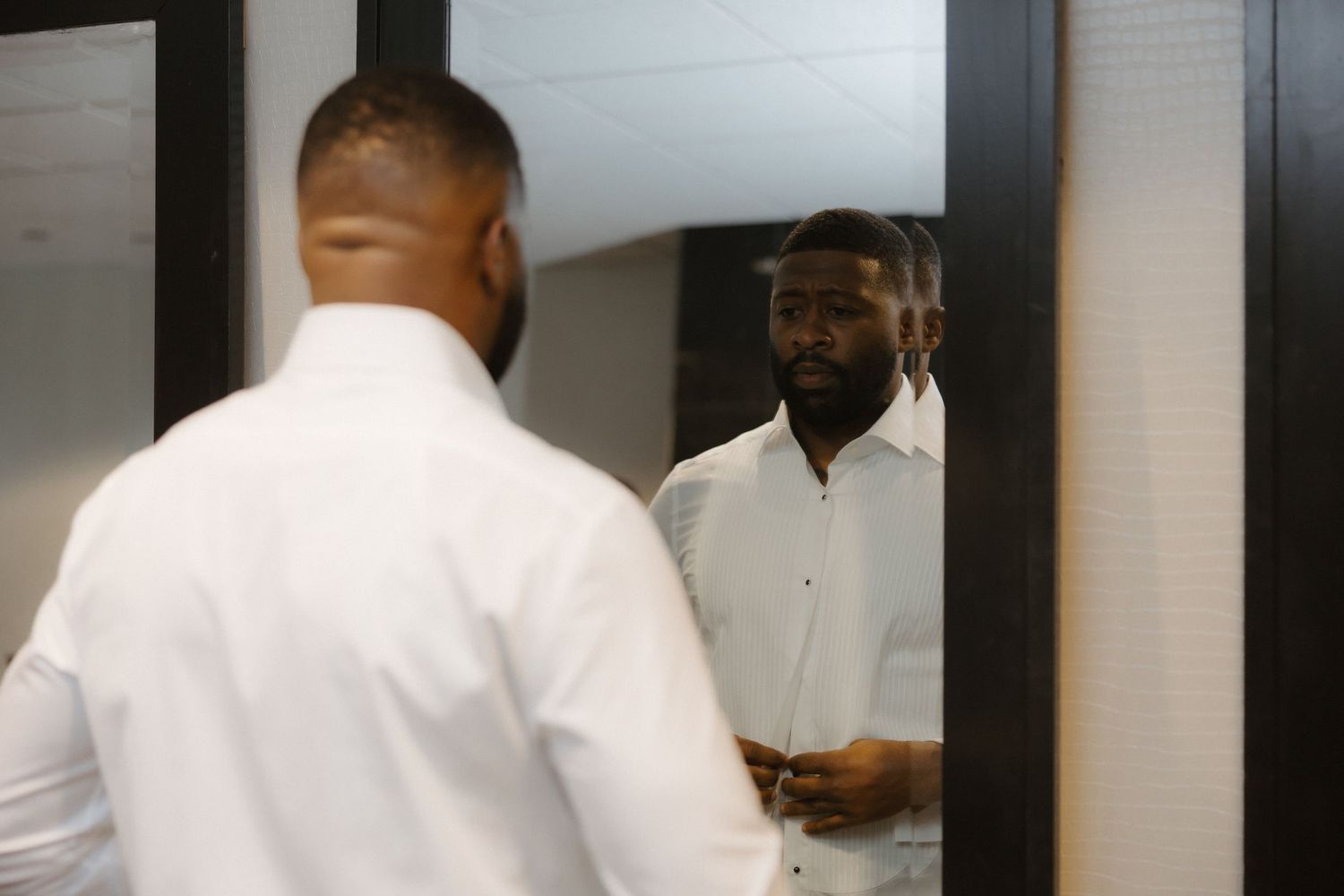 A man in a white dress shirt adjusts his outfit while looking at his reflection in a mirror.