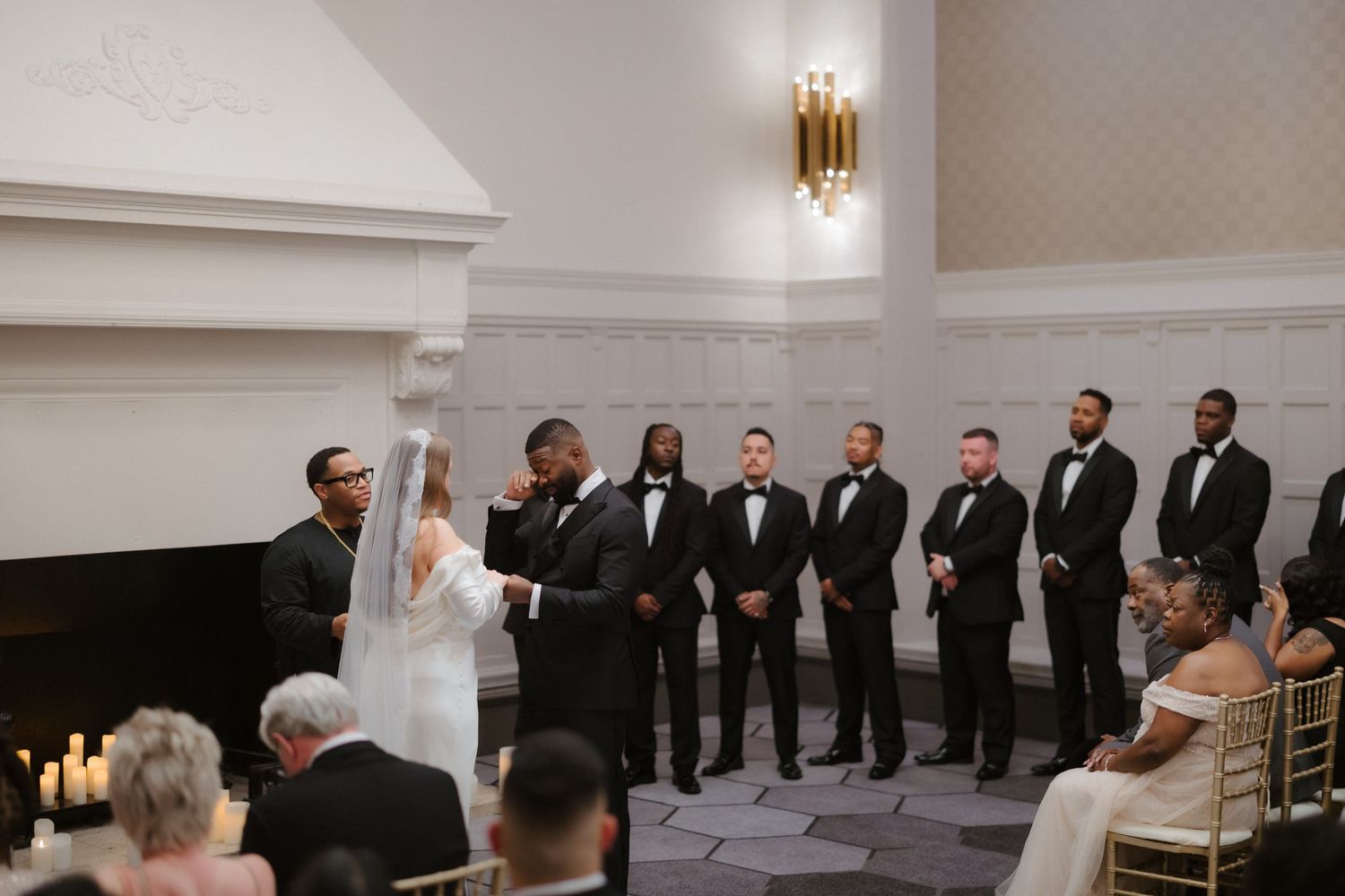Groom lifts bride's veil at elegant indoor wedding ceremony with groomsmen in black tuxedos standing nearby.