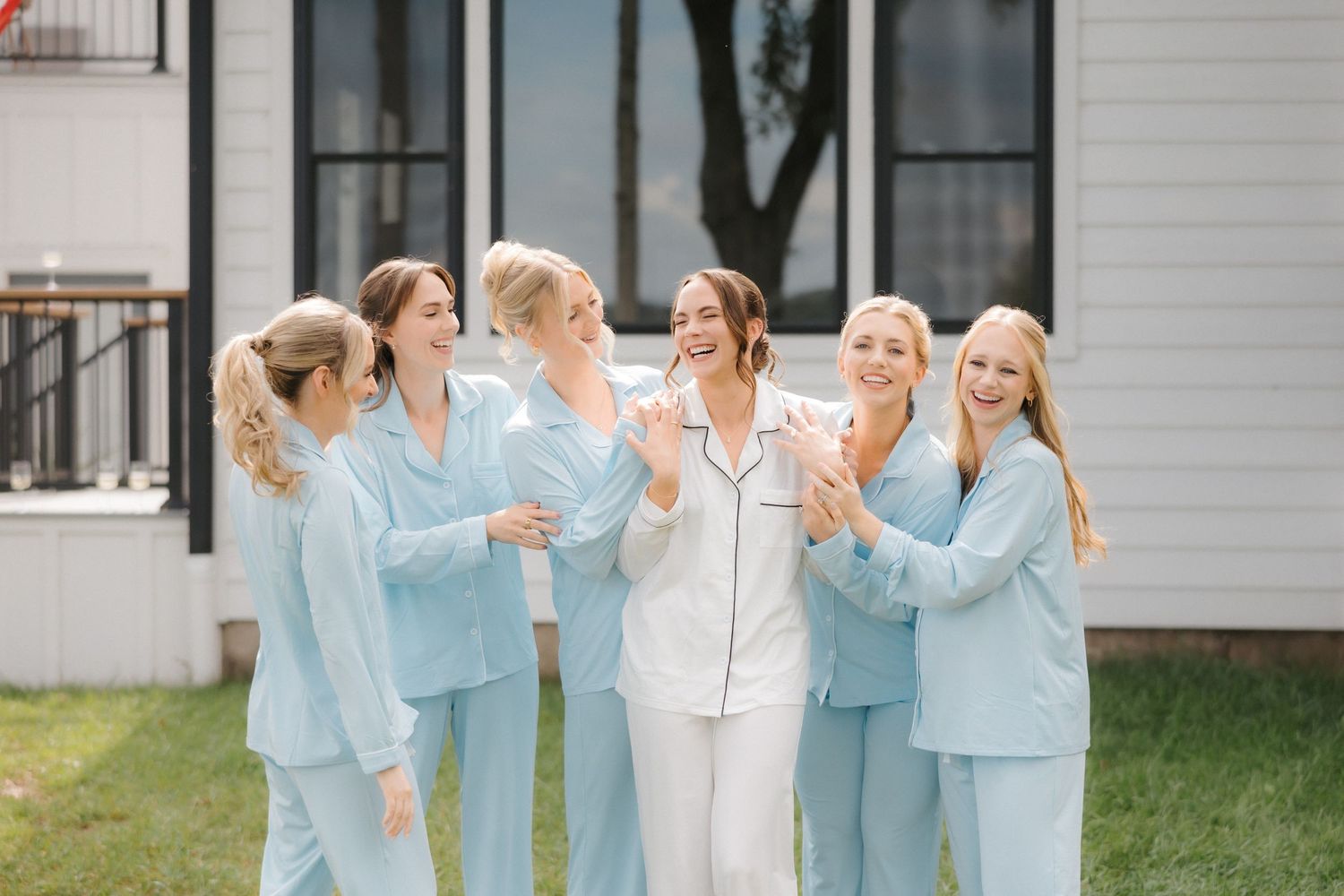 Smiling bridesmaids in light blue satin pajamas celebrate with bride in white pajamas outside on wedding day.