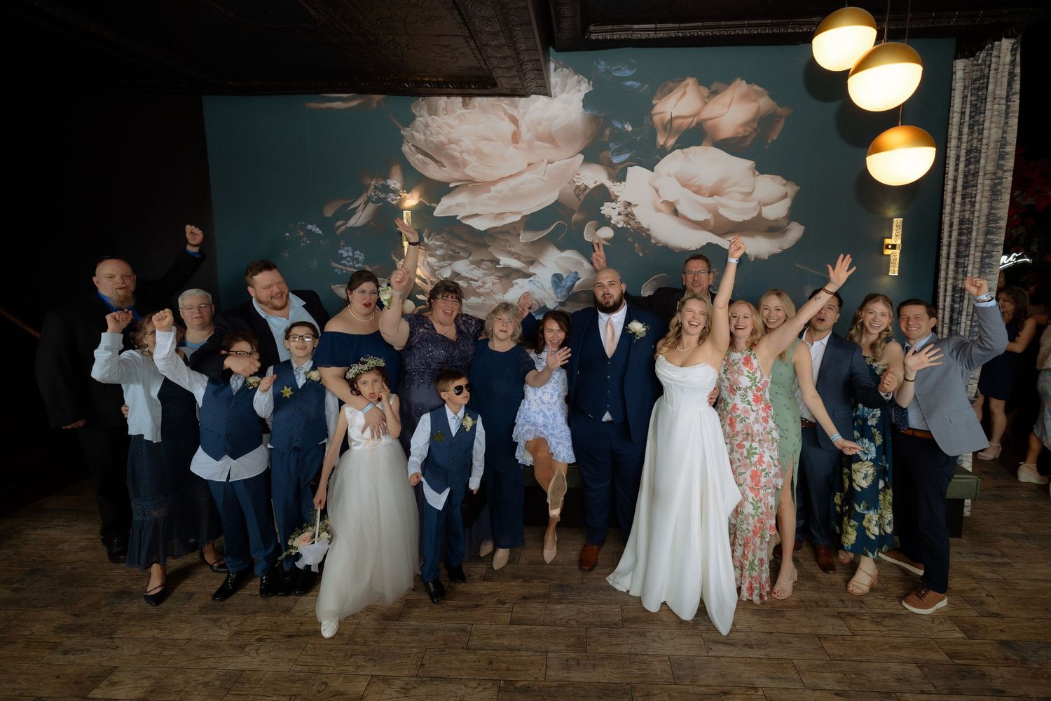 Wedding group photo with bride and groom surrounded by family and friends celebrating in front of floral mural.