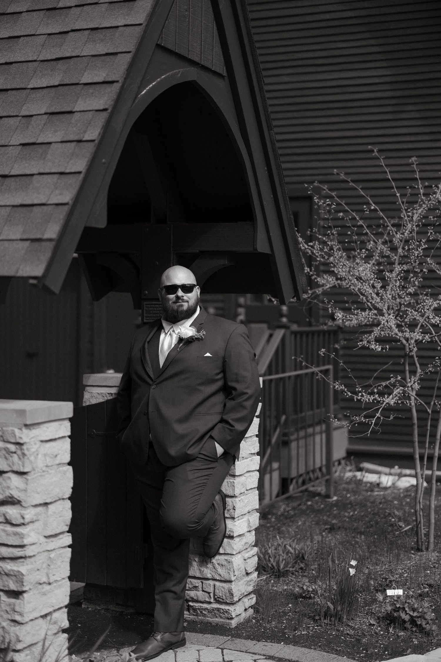 A well-dressed man in a suit and sunglasses leans against a stone pillar outside a chapel in a black and white photo.