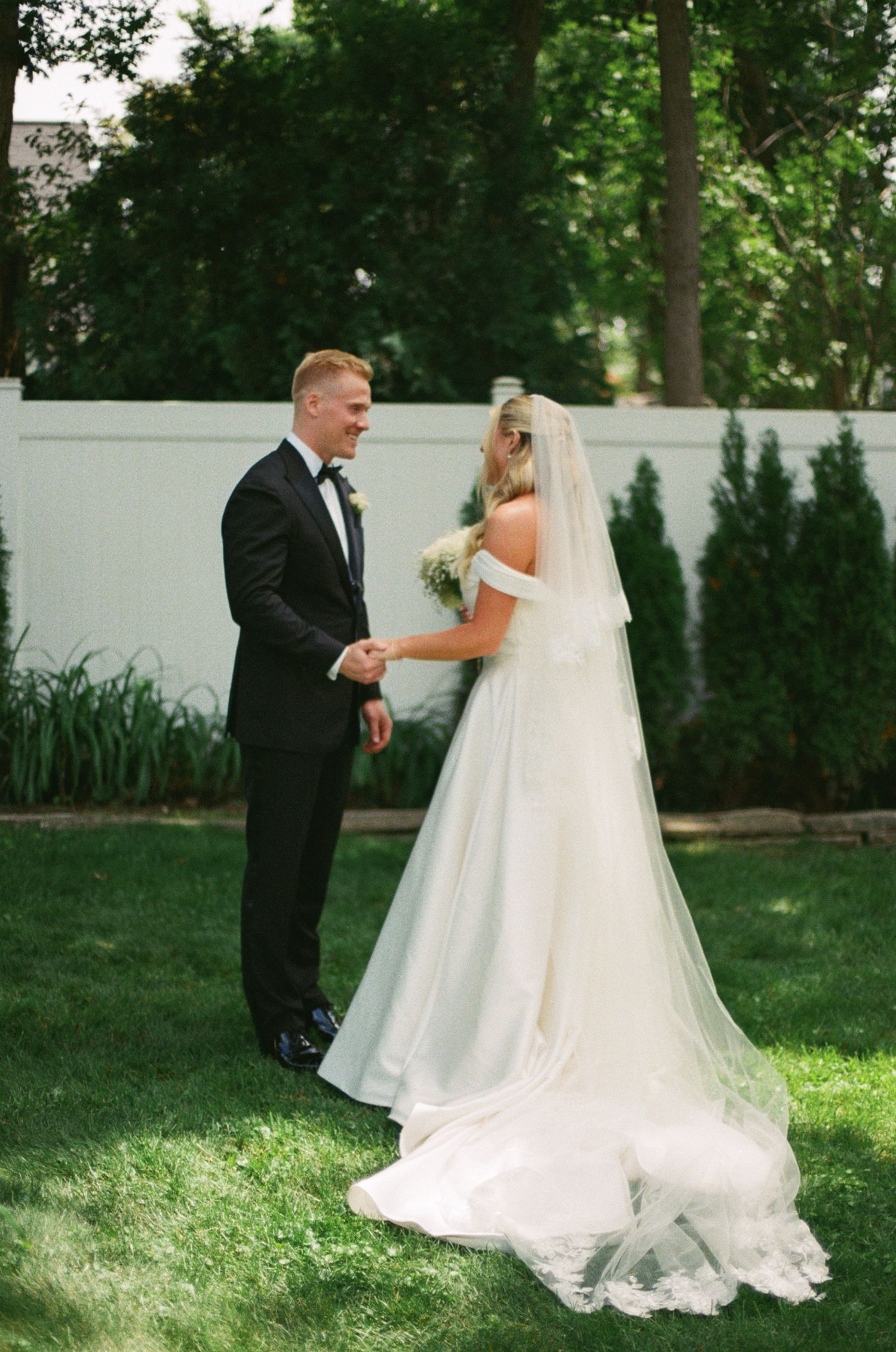 A smiling groom in a black tuxedo holds hands with his bride in a white off-shoulder gown and veil on a green lawn.