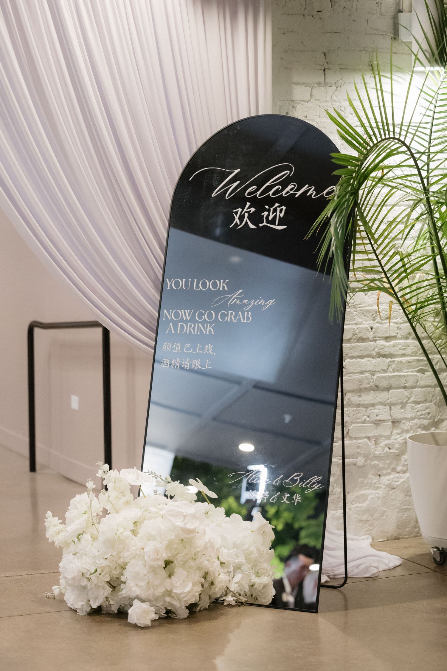 Elegant bilingual wedding welcome mirror sign with white floral arrangement, white calligraphy, and Chinese characters on dark background.