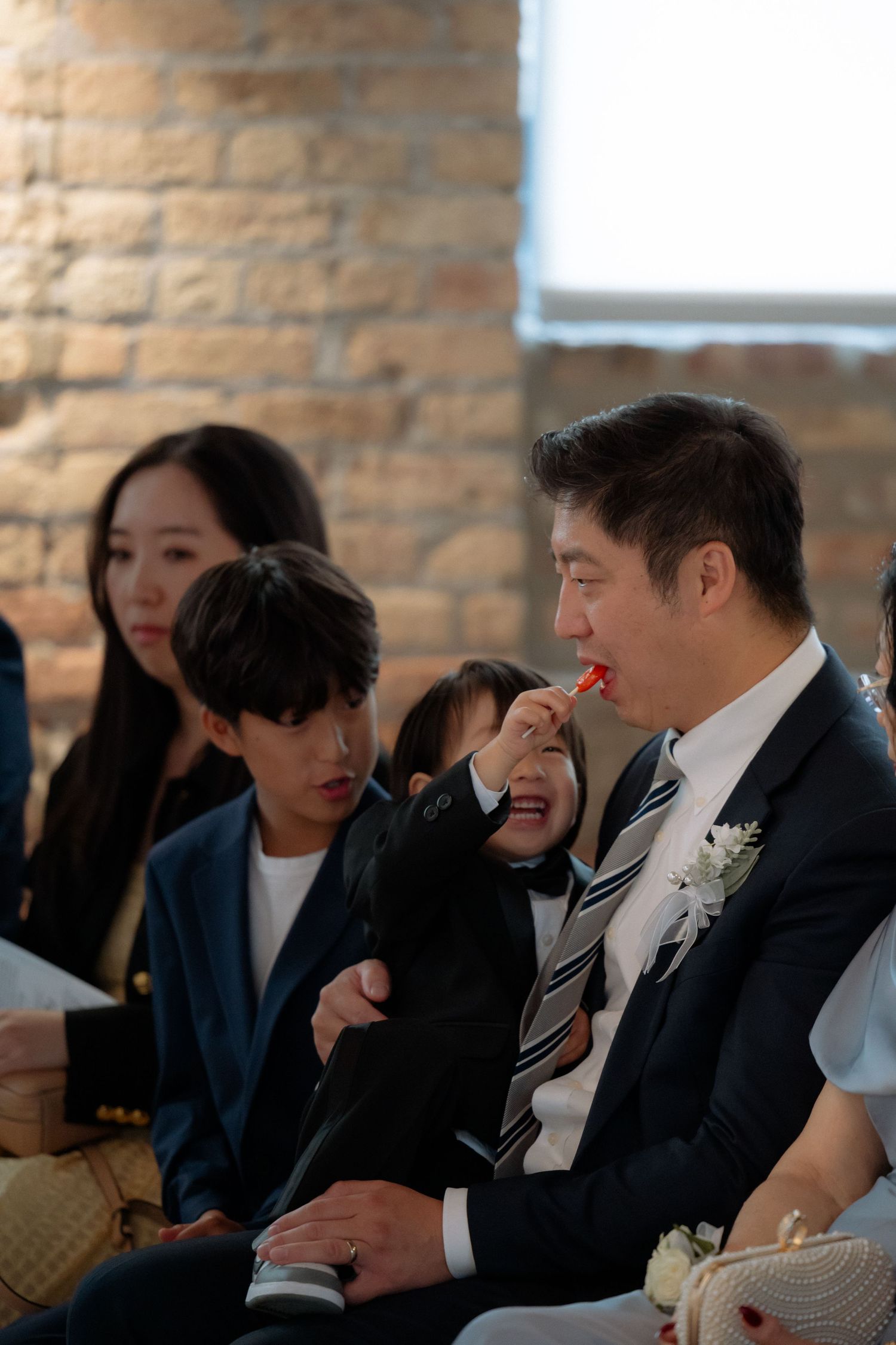 A man in a suit with a boutonniere feeds a child candy during a wedding ceremony while guests sit nearby.