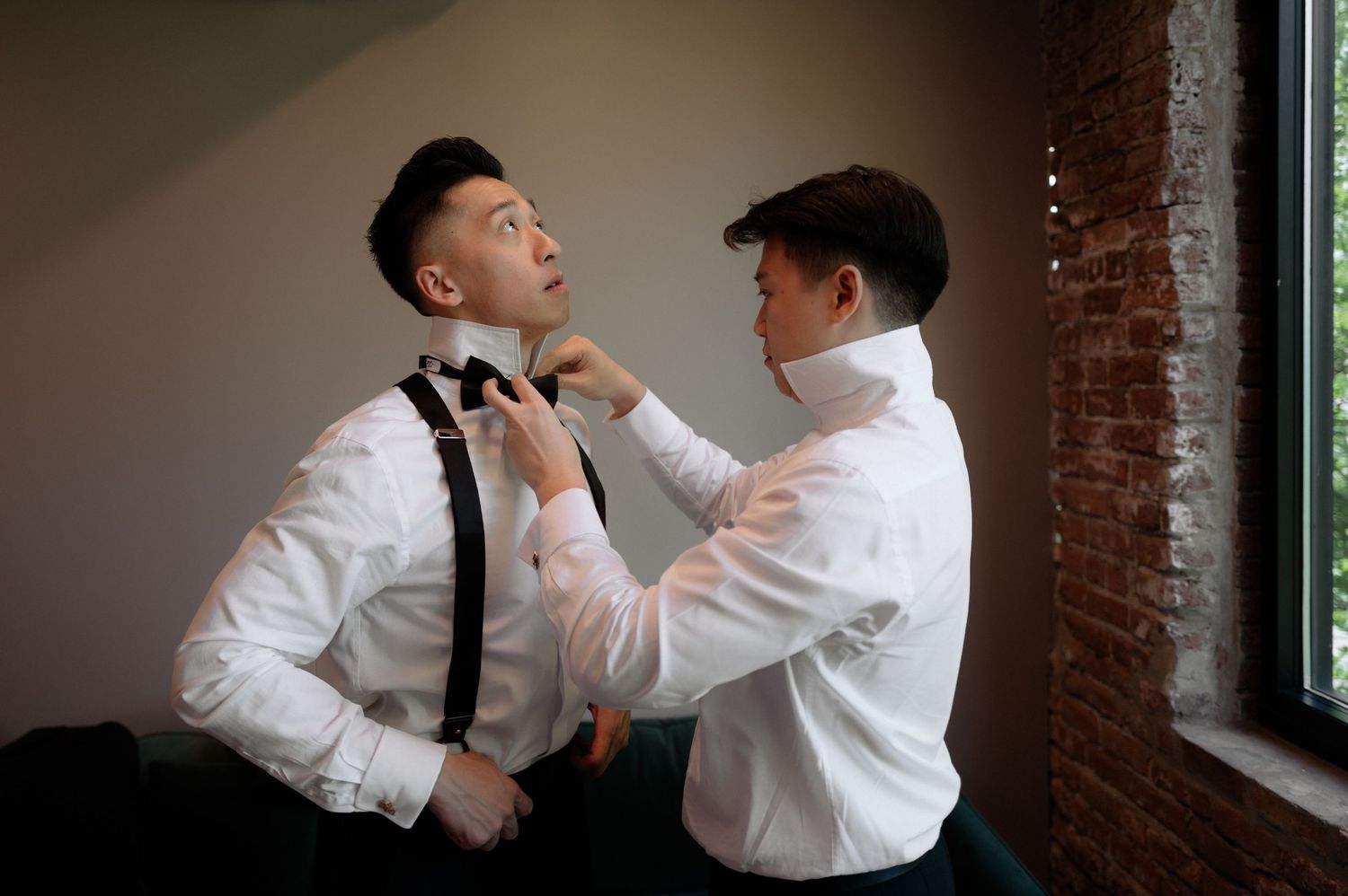 Two men in white dress shirts, one adjusting the other's black bow tie, preparing for a formal event.