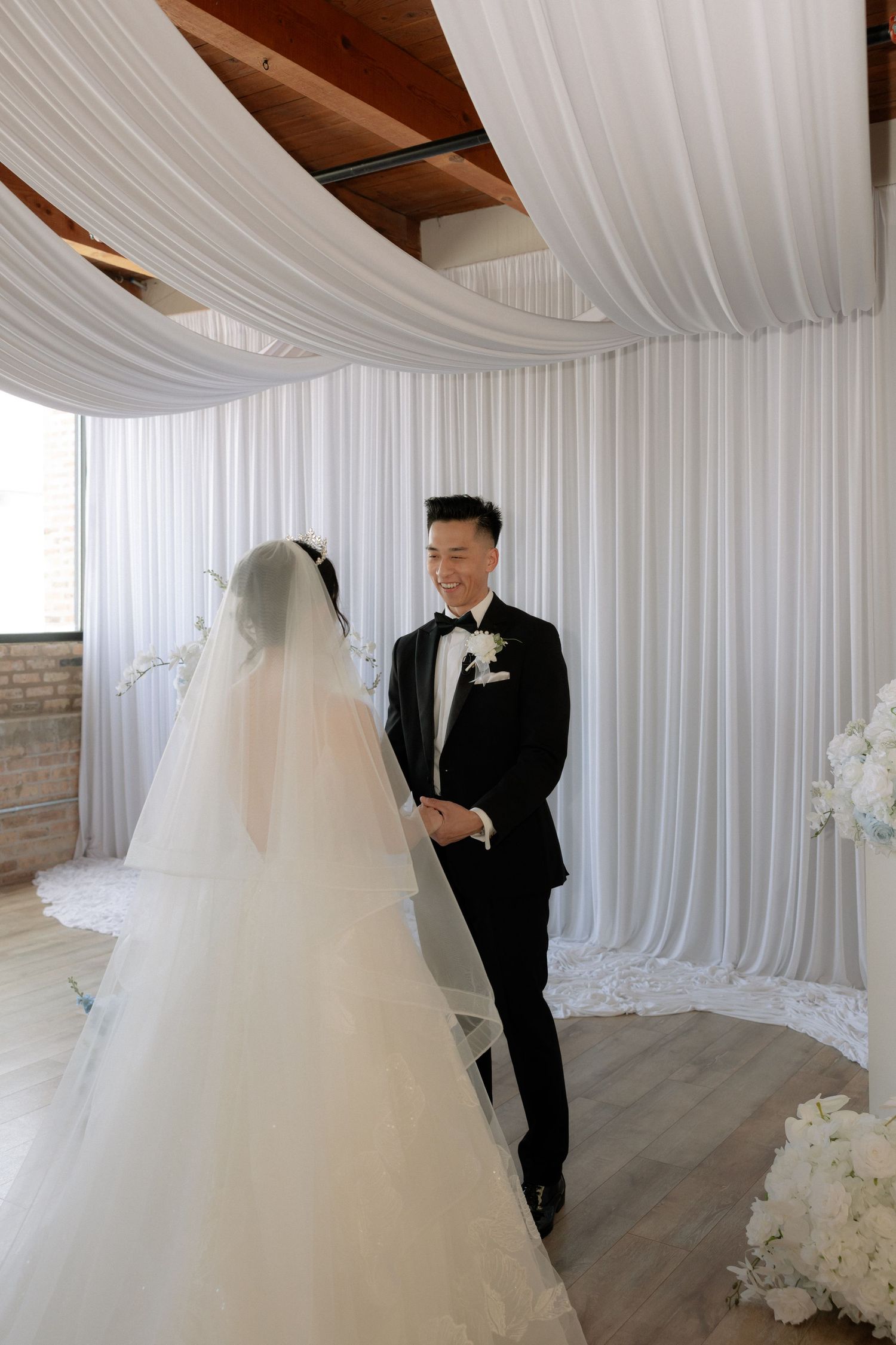 Groom in black tuxedo smiles at bride in white gown during indoor wedding ceremony with draped white backdrop.
