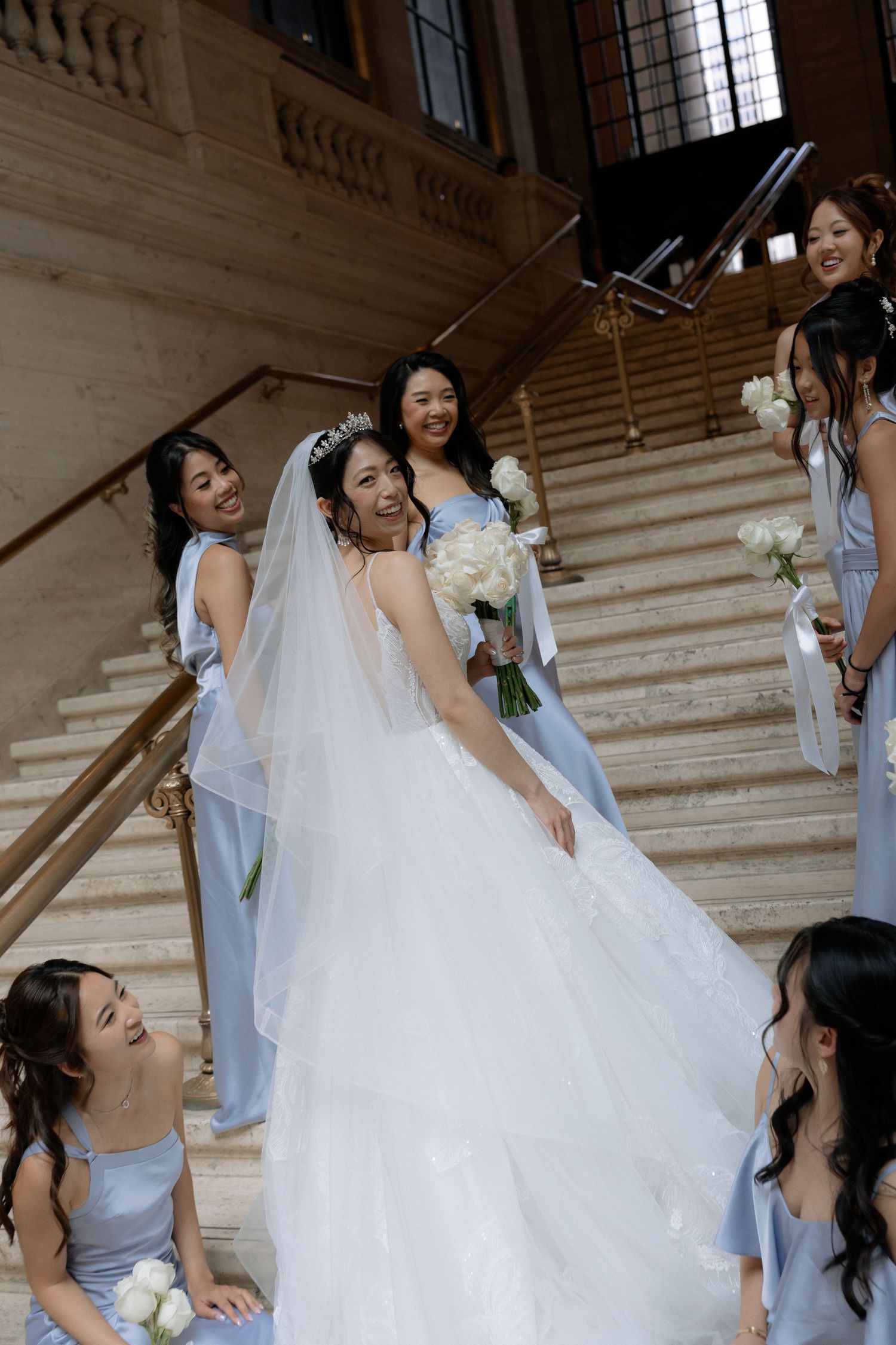 Smiling Asian bride in layered white gown with bridesmaids in light blue dresses on staircase.
