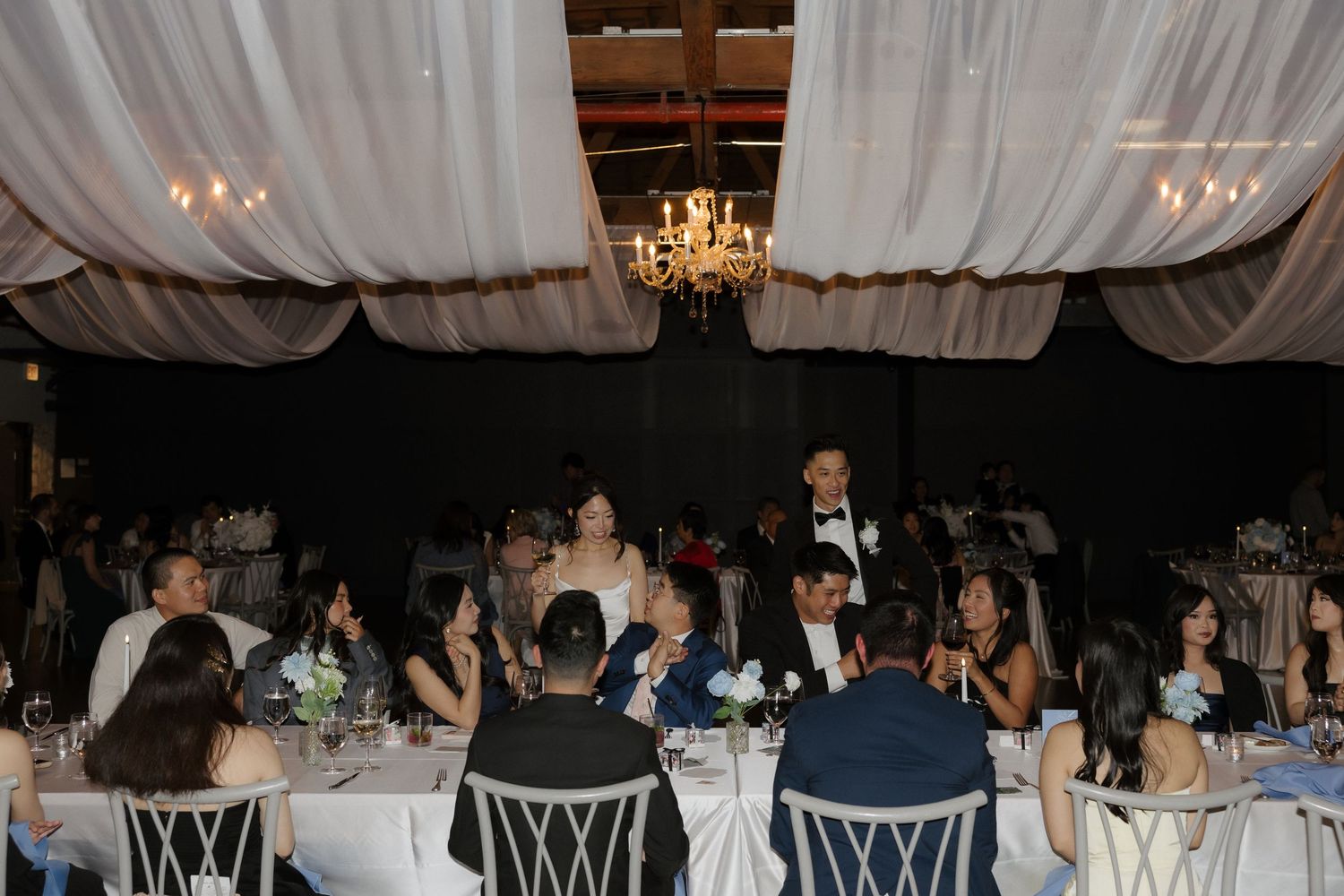 Elegant wedding reception in a decorated venue with chandelier lighting, guests seated at white tables with floral centerpieces.