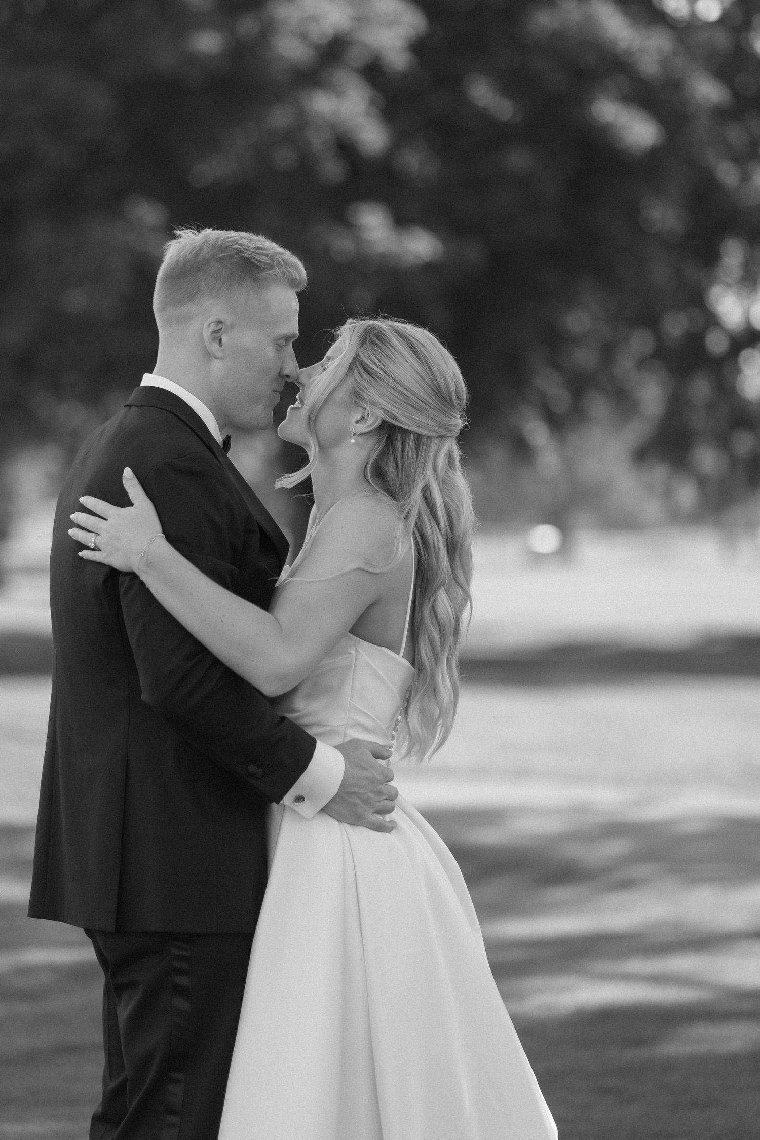 Black and white romantic portrait of bride and groom embracing closely outdoors under trees.