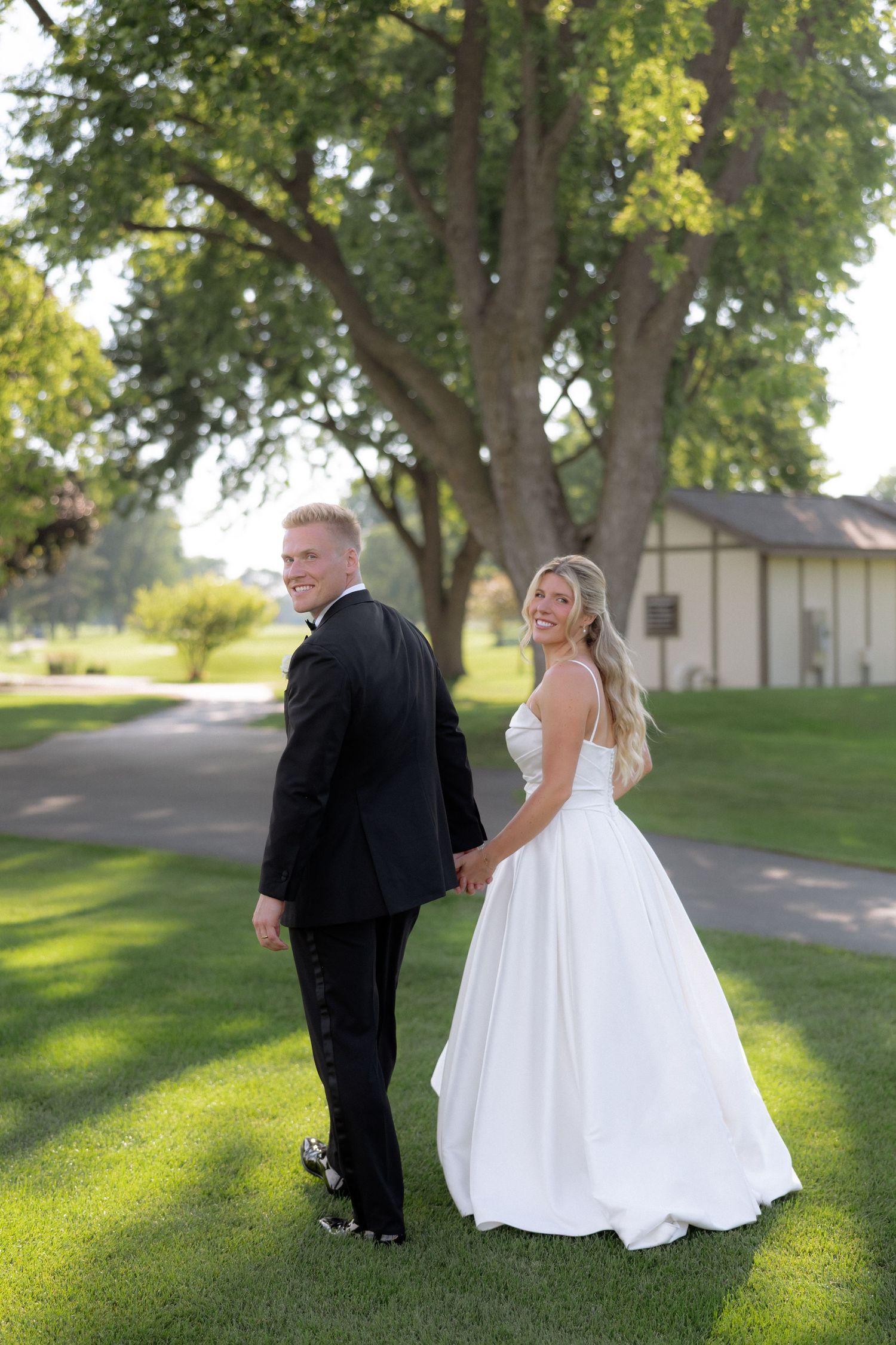 Bride and groom holding hands walking on green lawn, smiling back at camera with large trees behind them.