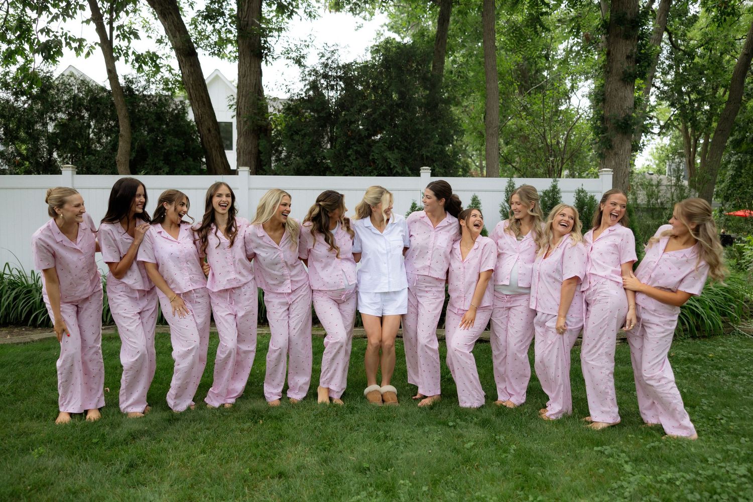 Bride in white surrounded by bridesmaids wearing matching pink pajamas, posing together on green lawn.