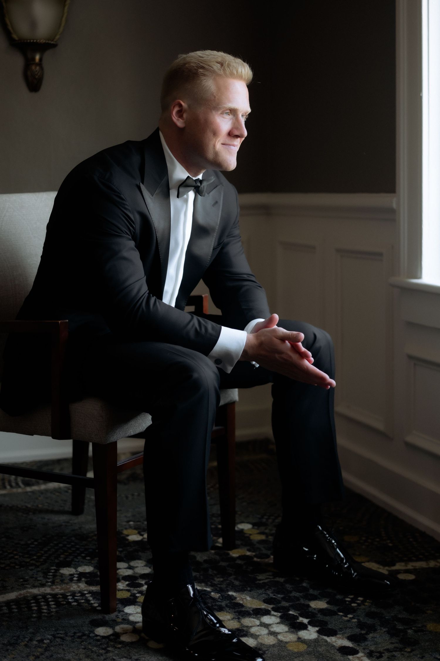 Groom in black tuxedo sitting in chair gazing out window before wedding ceremony.